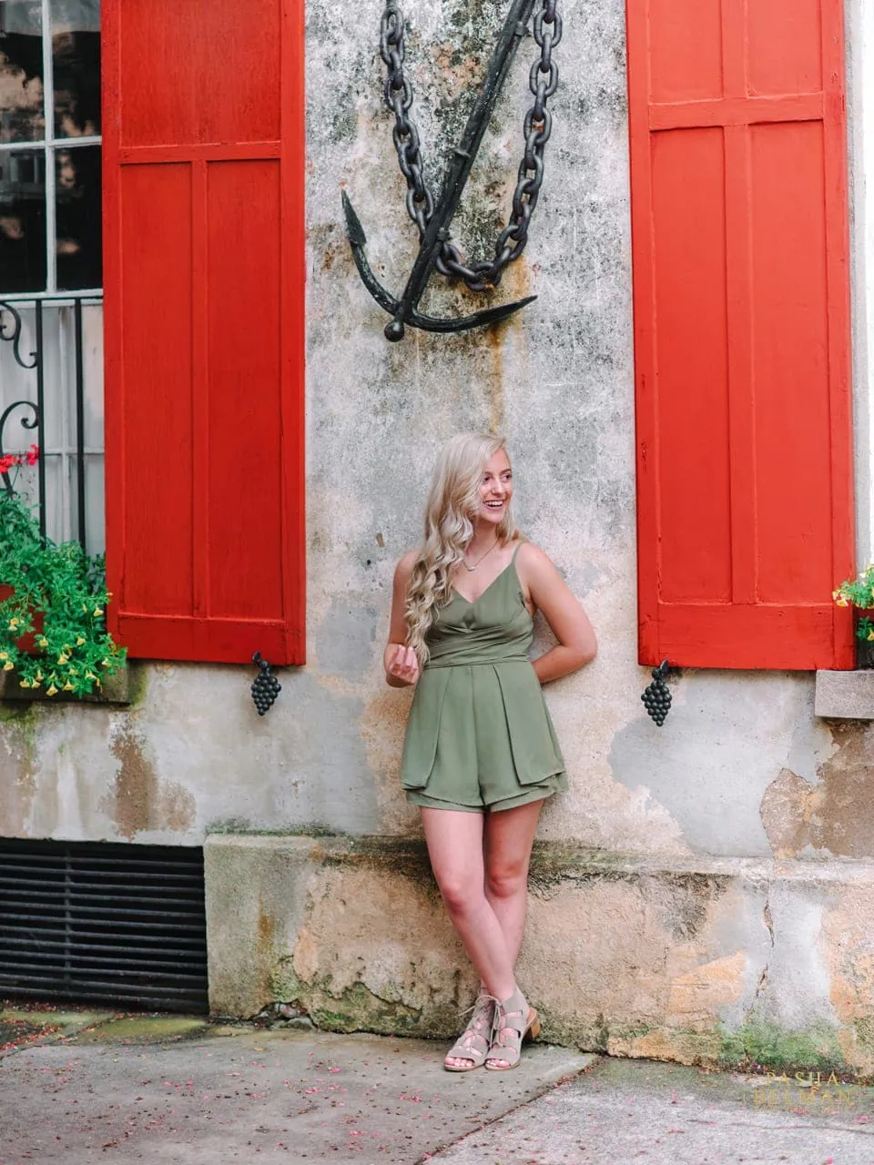 Blonde senior girl in olive green romper smiling beside red window shutters and iron anchor in historic Charleston