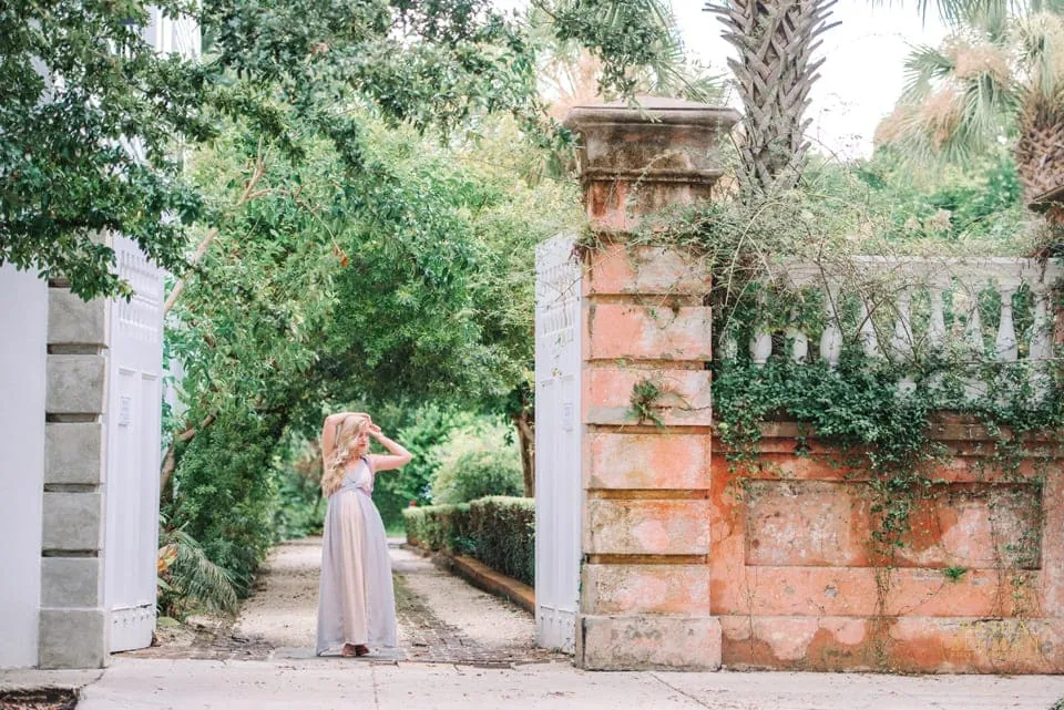 Senior girl wearing a floppy hat and flowy dress standing under arching greenery between historic Charleston walls