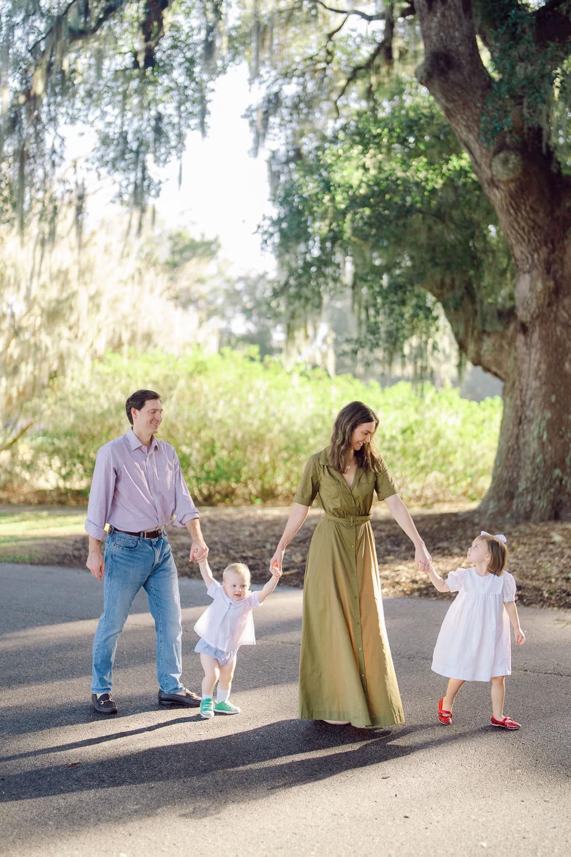 Family beach portrait featuring a happy family with young children walking hand-in-hand on a sunny day near a large oak tree, capturing candid moments and natural light.