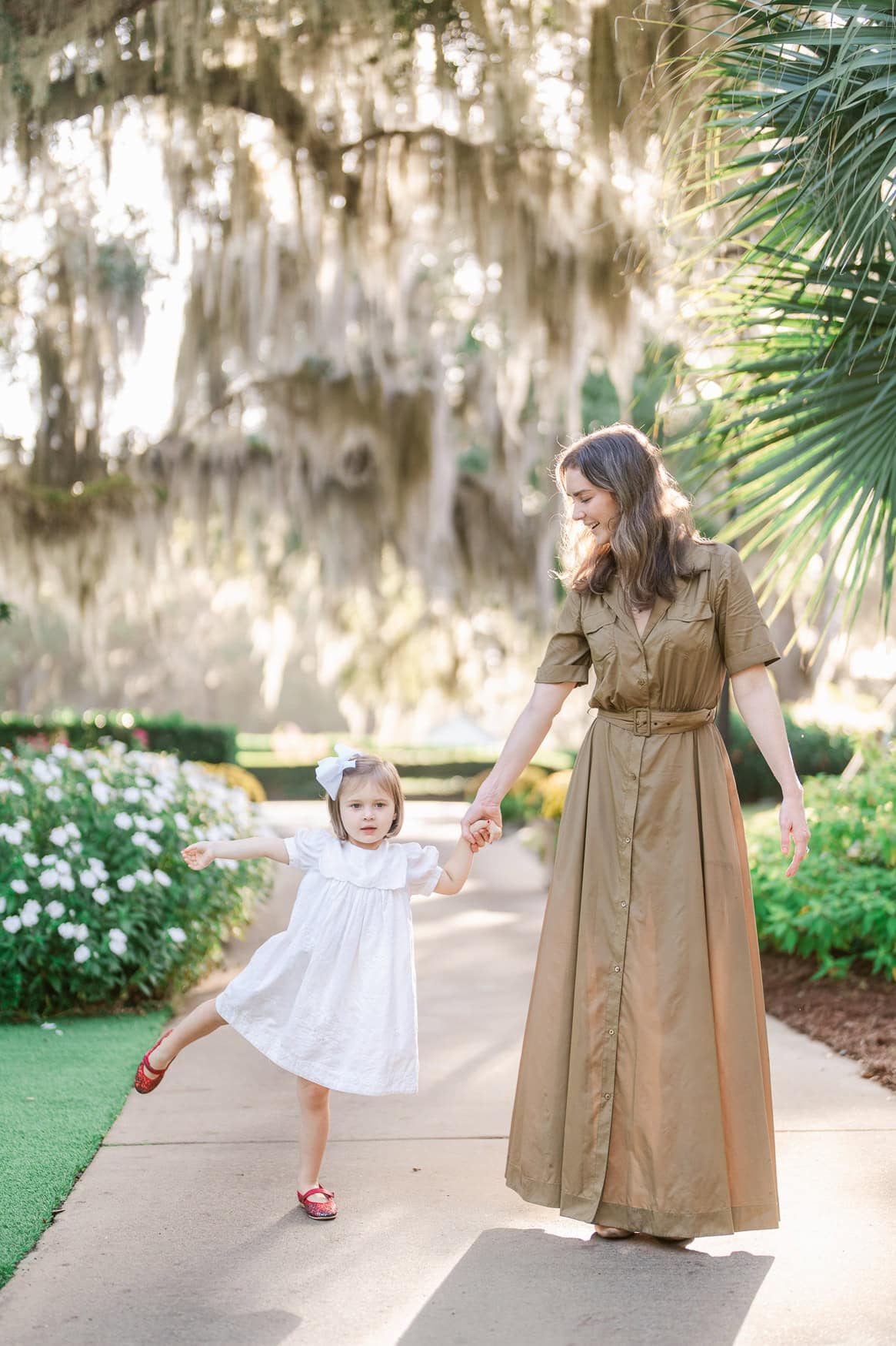 Baby girl twirling in a white dress with mother holding her hand at a park with Spanish moss trees, captured during golden hour, creating a warm and joyful family portrait.