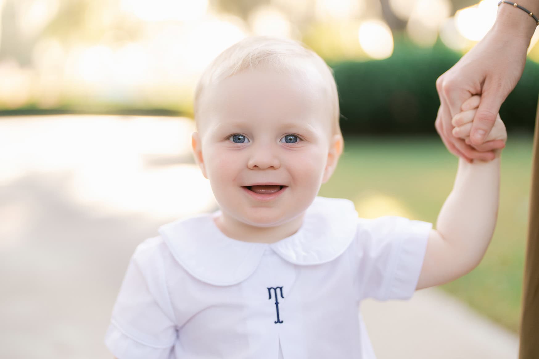 Cute toddler girl with bright blue eyes and blonde hair smiling outdoors during a family photoshoot, holding an adult's hand, in a natural park setting.