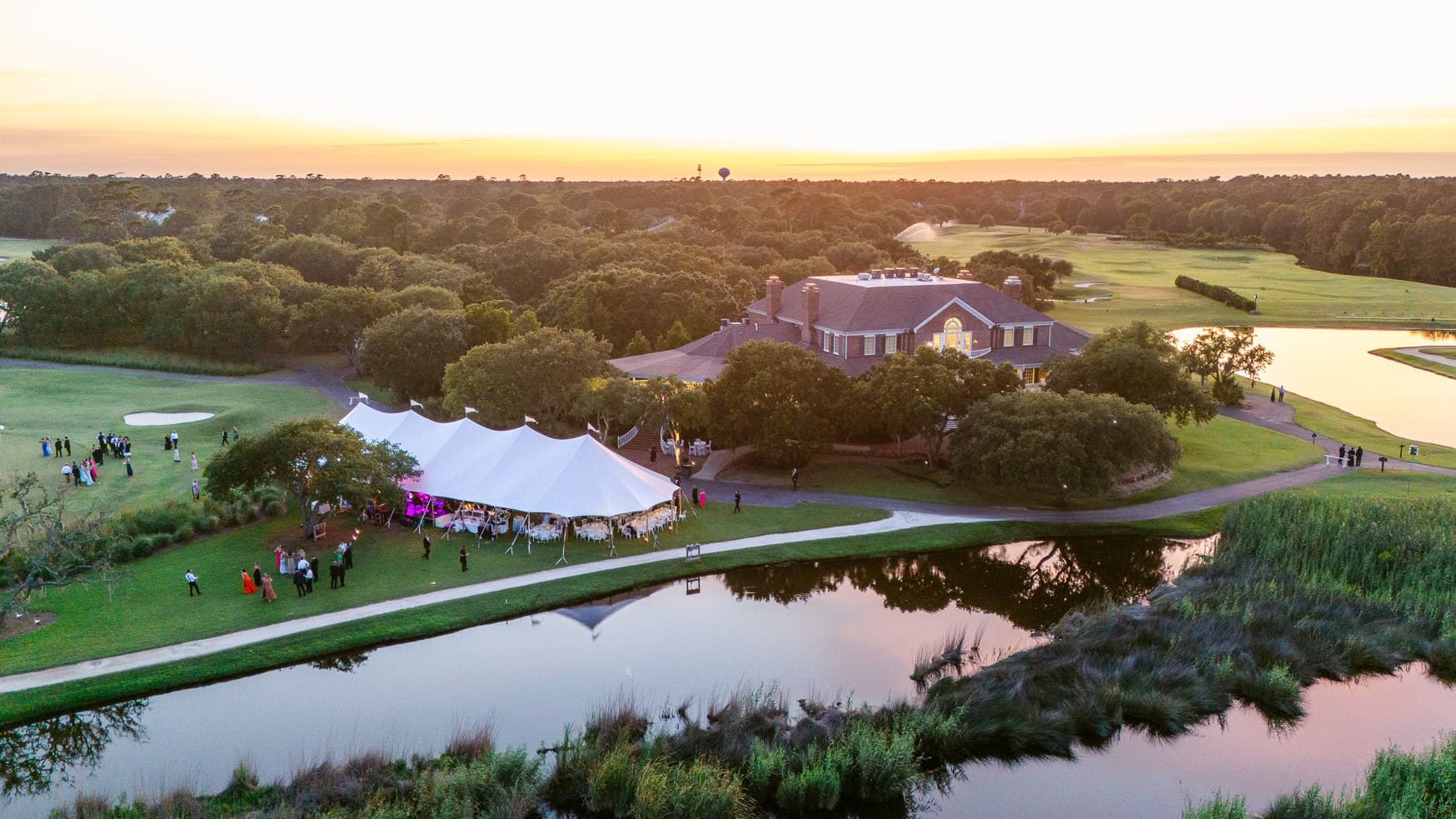 Vibrant sunset over a wedding reception outdoors at a picturesque golf course with a pavilion, surrounded by lush greenery and water features, capturing elegant wedding celebration photos.