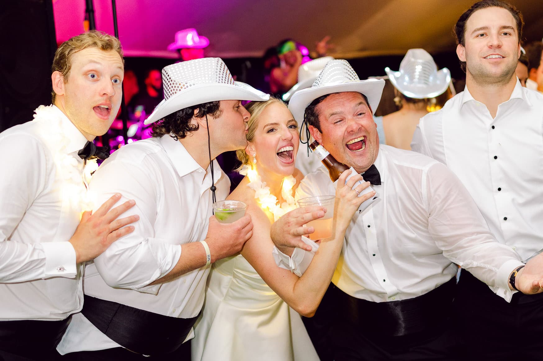 Festive wedding guests wearing cowboy hats celebrate and dance at a lively wedding reception with drinks and joyful expressions.