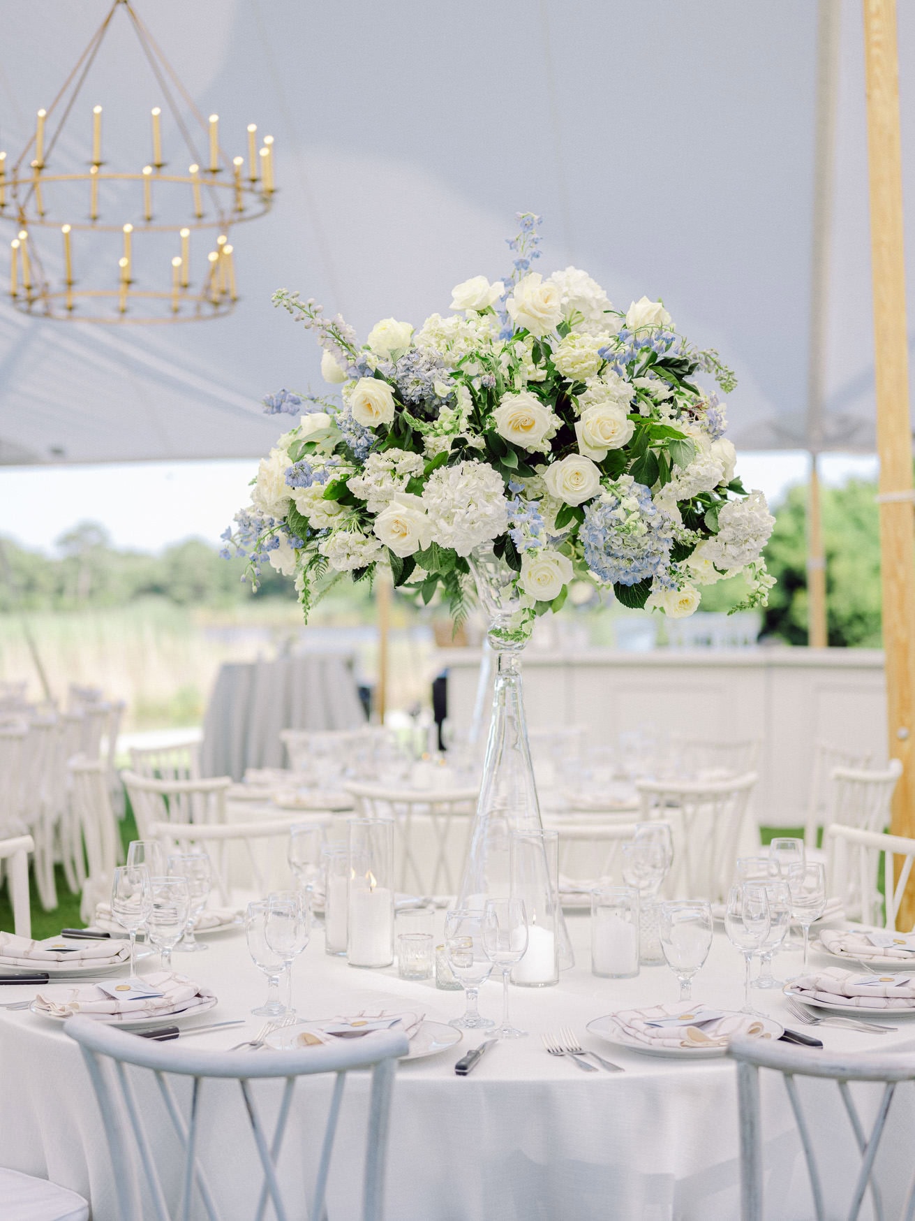Elegant wedding reception table setup with a tall floral centerpiece of white roses and hydrangeas under a white canopy, perfect for outdoor wedding photography.