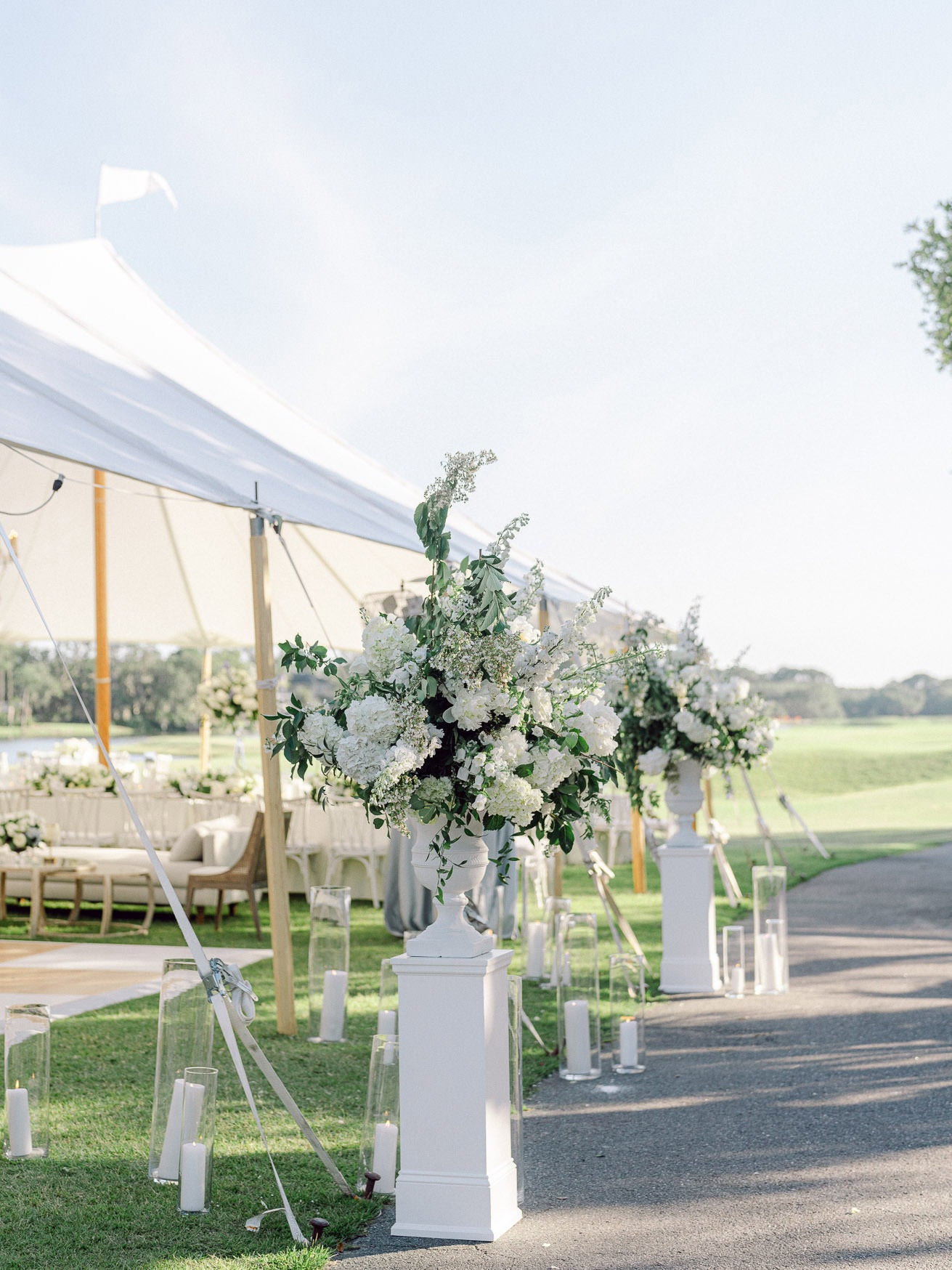 Luxurious white floral arrangements on elegant pedestals at an outdoor wedding ceremony setup with white tents and candle lanterns.