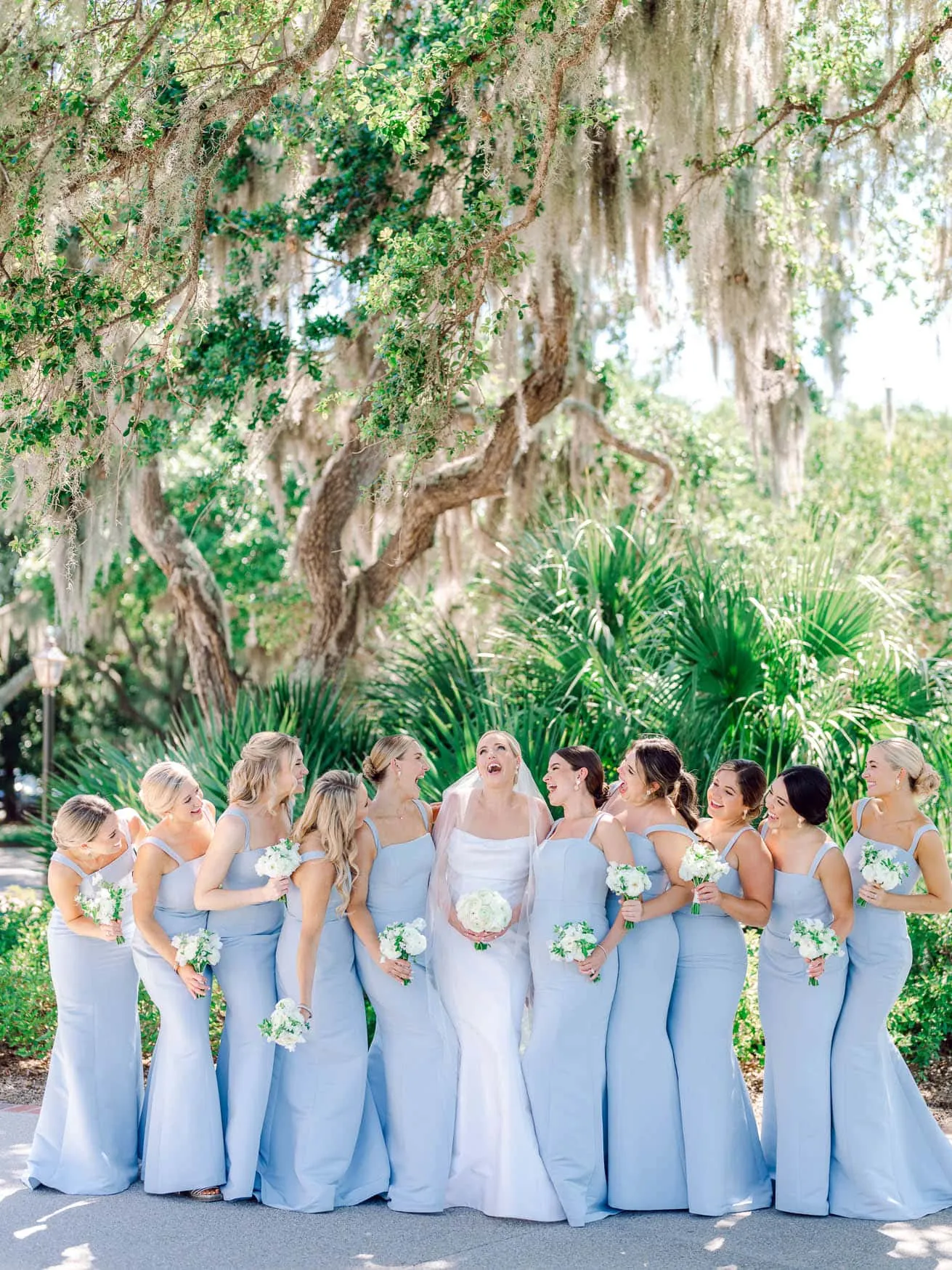 Beautiful bride and bridesmaids enjoying a joyful moment outdoors in a lush, green garden setting with hanging moss and tropical plants, perfect for wedding photography and family beach portraits.