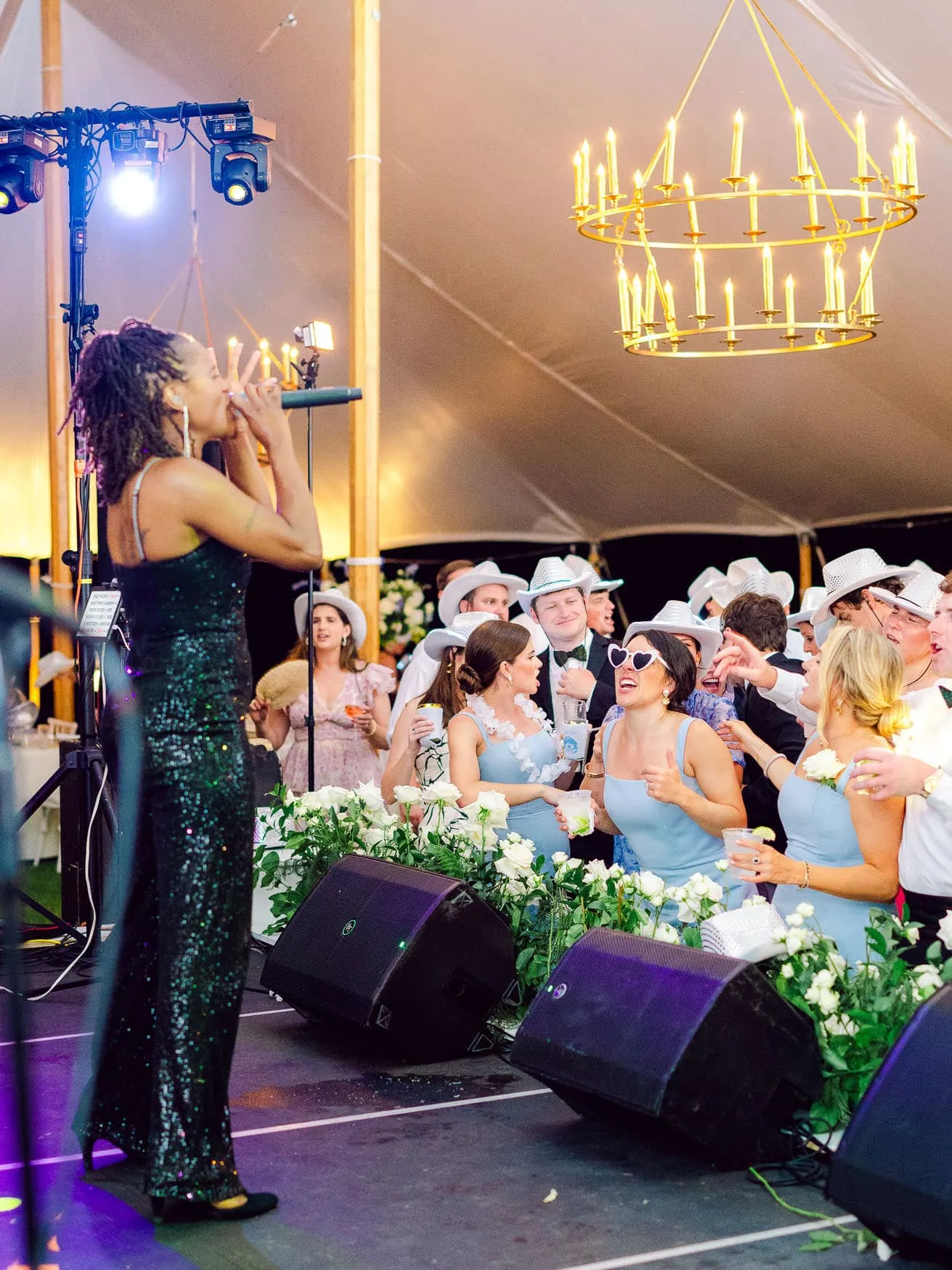 Beautiful female singer performing at wedding reception with guests dancing and celebrating under elegant chandelier in large wedding tent at sunset.
