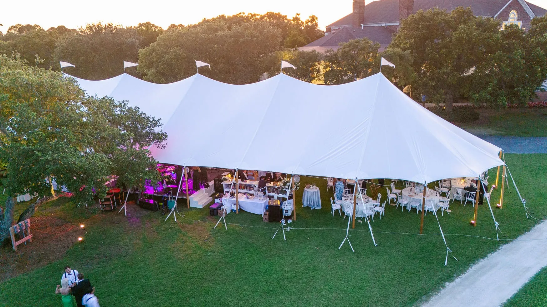 Beautiful outdoor wedding reception under a large white tent in the evening with elegant table settings and floral arrangements, surrounded by lush greenery and a serene landscape.