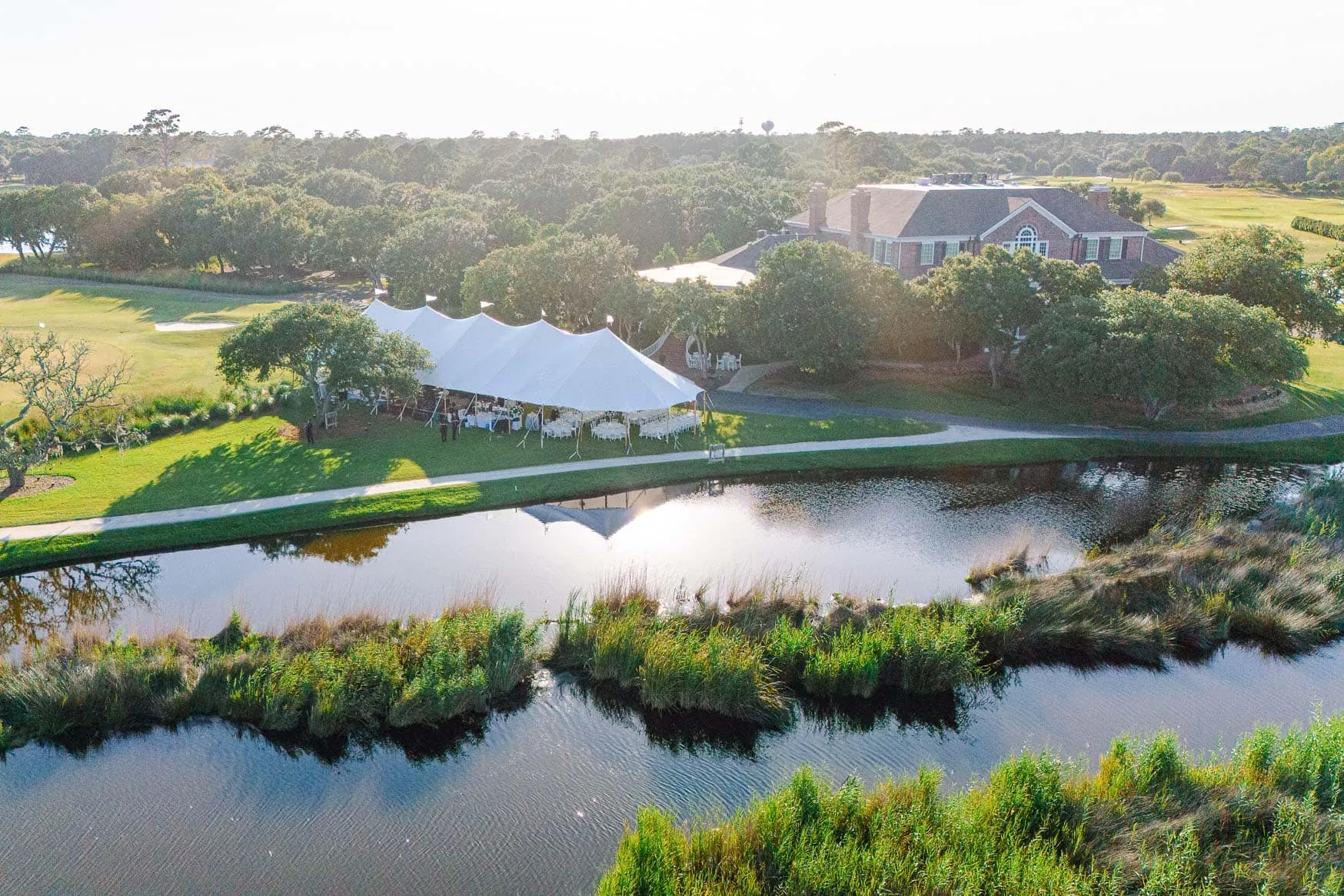 Beautiful outdoor wedding setup on a lush green golf course with a large white tent, water features, and surrounding trees, ideal for wedding photography and special events.
