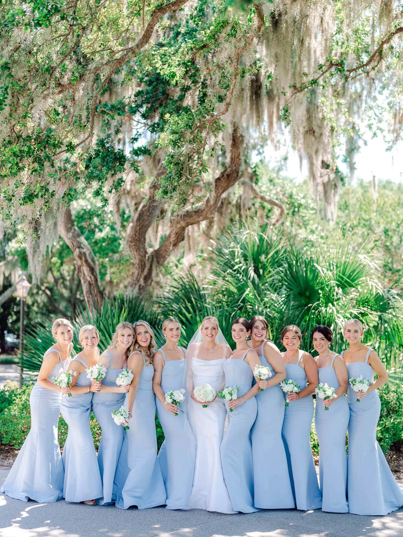 Bright bridal party in matching light blue dresses outdoors in front of lush greenery and large trees with hanging moss, capturing a joyful wedding moment in natural sunlight.
