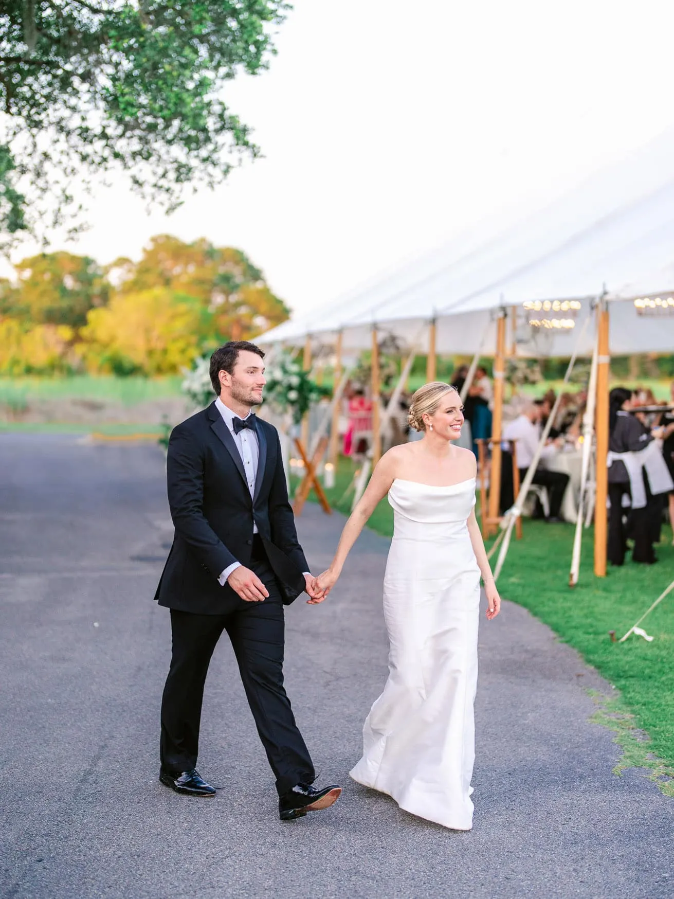 Elegant bride and groom holding hands at outdoor wedding reception during golden hour, with scenic natural background and stylish wedding attire.