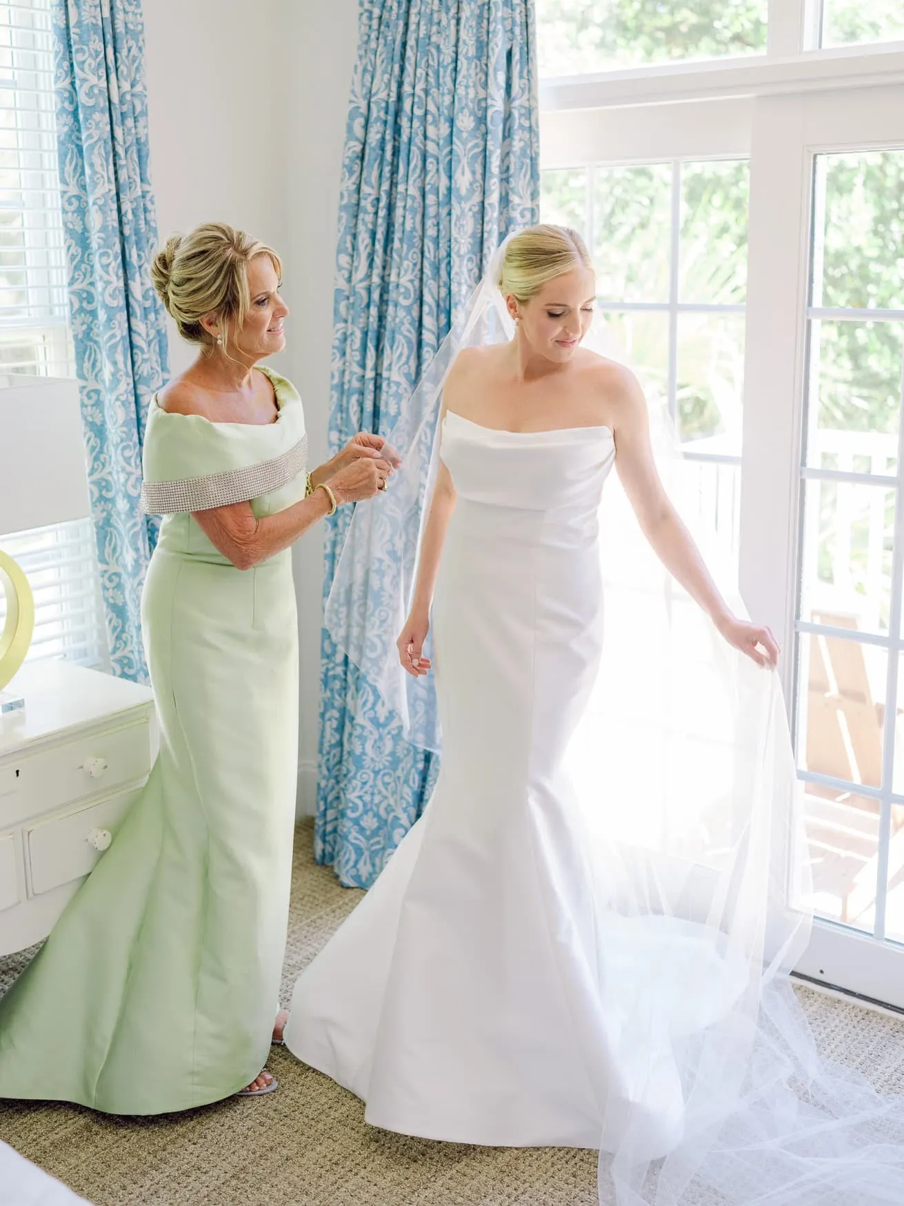 Elegant bride getting ready in bright room with her mother, capturing special wedding moments, beautiful white wedding gown, indoor wedding preparation, joyful family wedding day.
