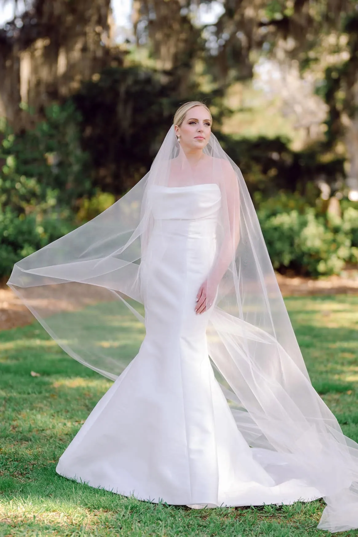 Elegant bride in a strapless white wedding gown with a long veil, standing outdoors in a lush green park, capturing timeless wedding photography moments.