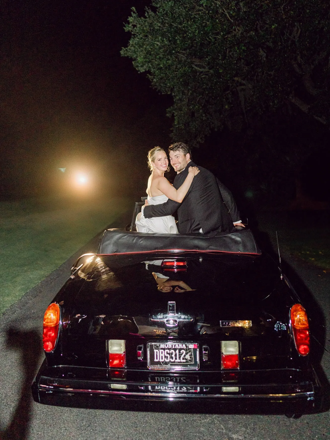 Elegant newlyweds sitting in a classic black convertible car during nighttime wedding photoshoot with moonlit background.