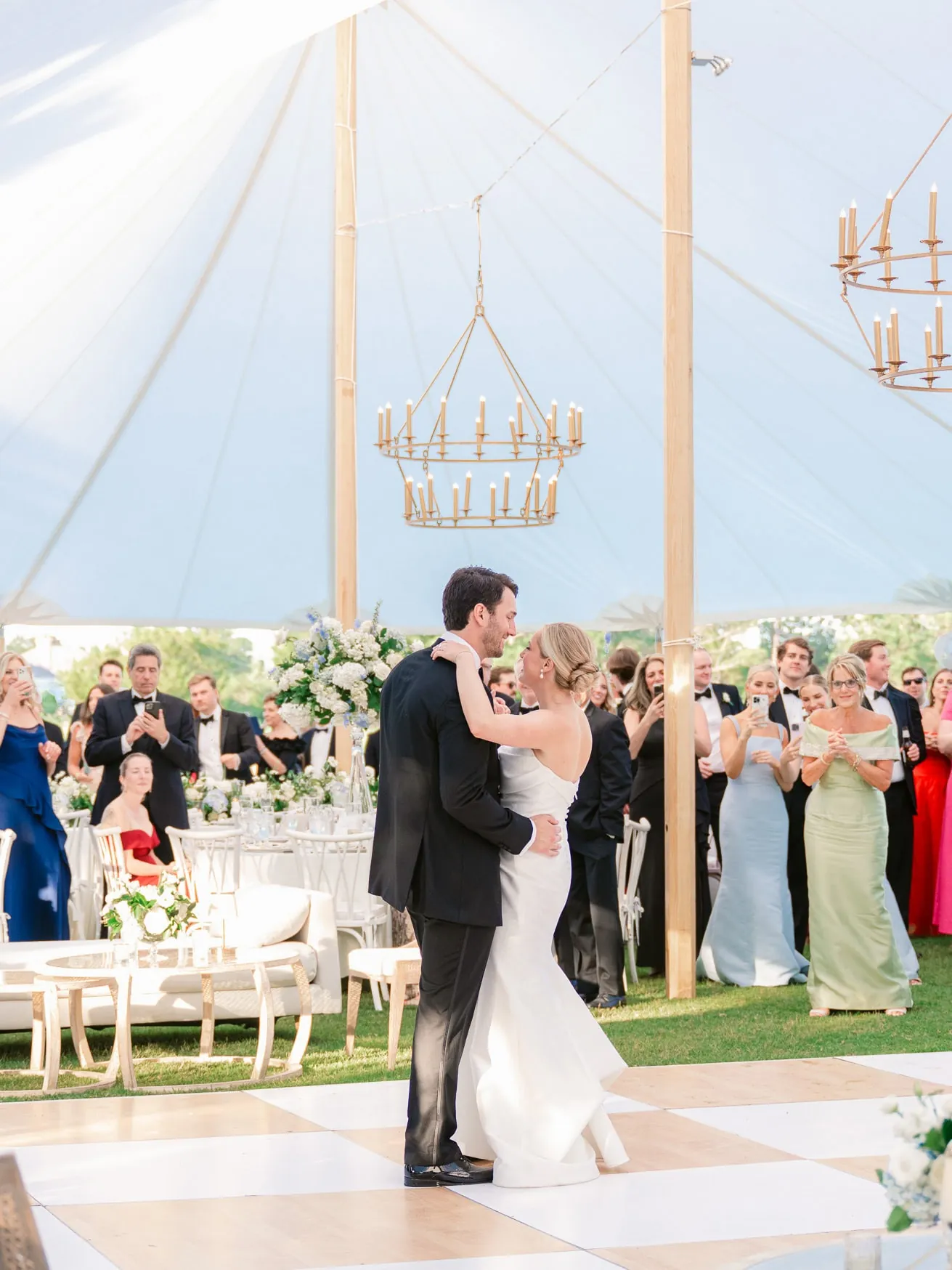 Elegant outdoor wedding couple sharing their first dance under a white tent with chandeliers, surrounded by guests, floral arrangements, and a lush green lawn, capturing a romantic wedding moment.
