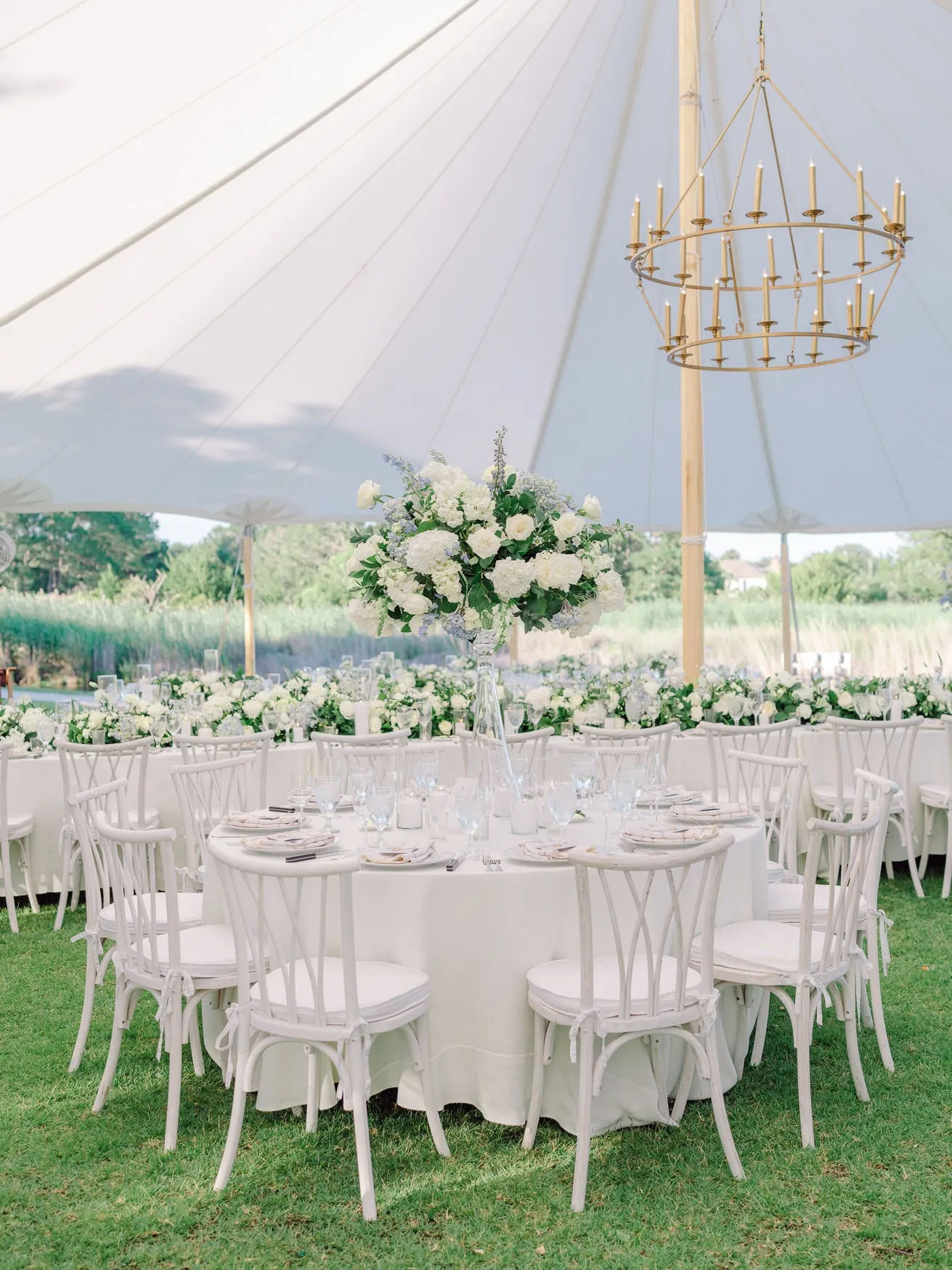 Elegant outdoor wedding reception setup with white floral centerpiece, chic tableware, and decorative chandelier under a white canopy on lush green grass.
