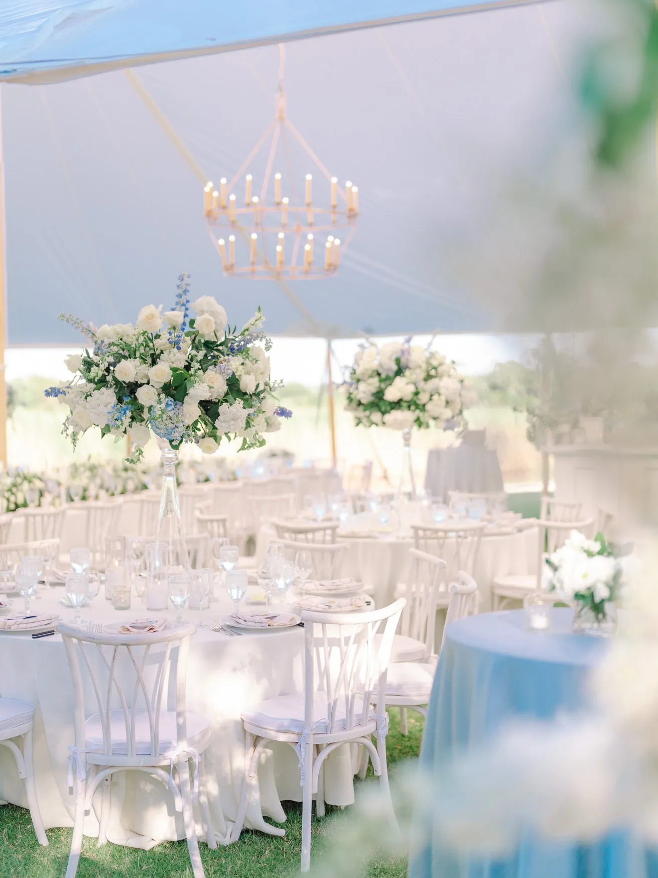 Elegant outdoor wedding reception table setup adorned with tall floral centerpieces of white roses and greenery, under a blue canopy with a modern chandelier, perfect for romantic wedding photography.
