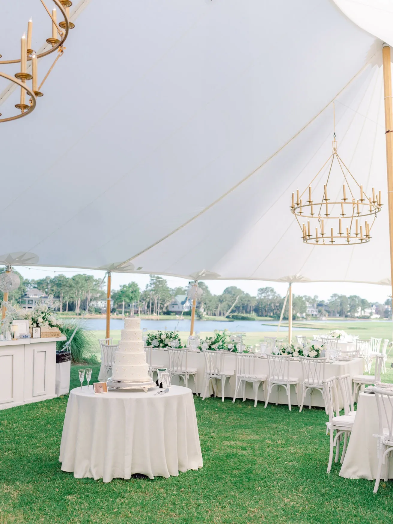 Elegant outdoor wedding reception under a large white tent with a tiered wedding cake on a round table, surrounded by white chairs and lush greenery, overlooking a tranquil lake.