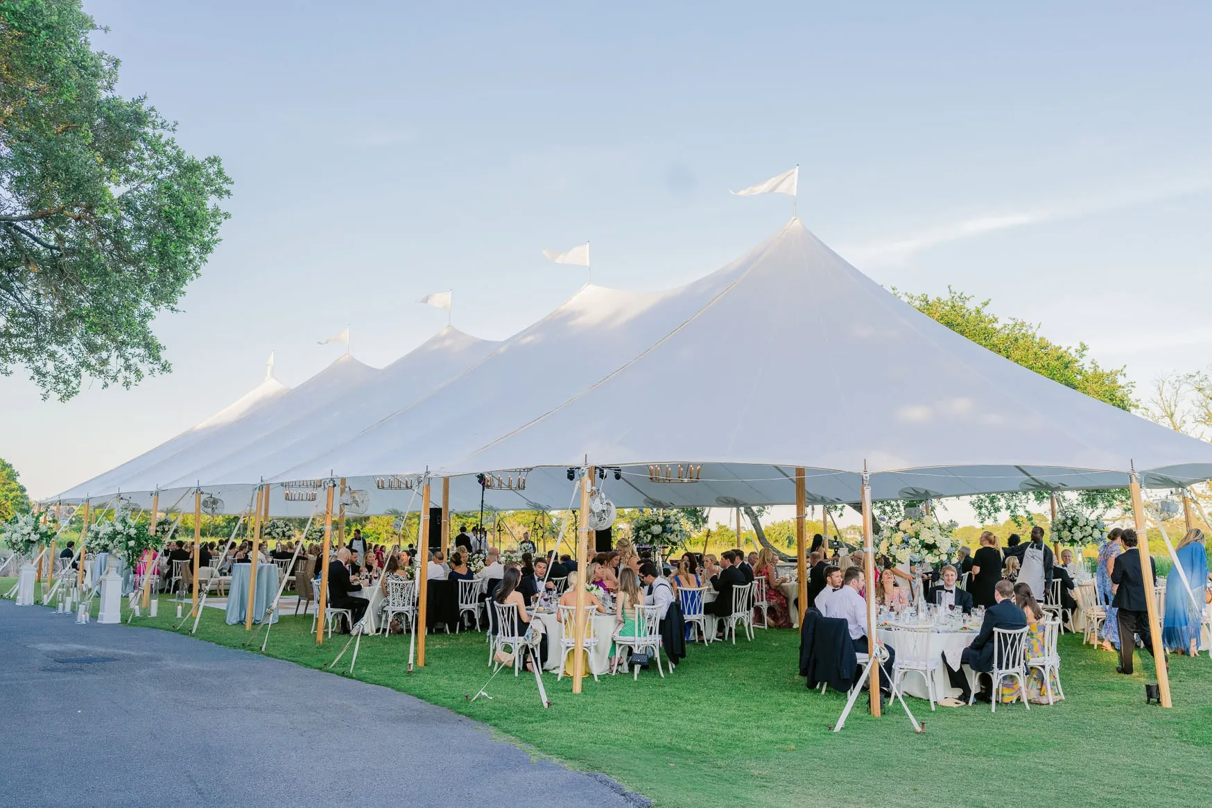 Elegant outdoor wedding under a large white tent with guests seated at decorated tables, surrounded by lush greenery and floral arrangements, captured during daytime.