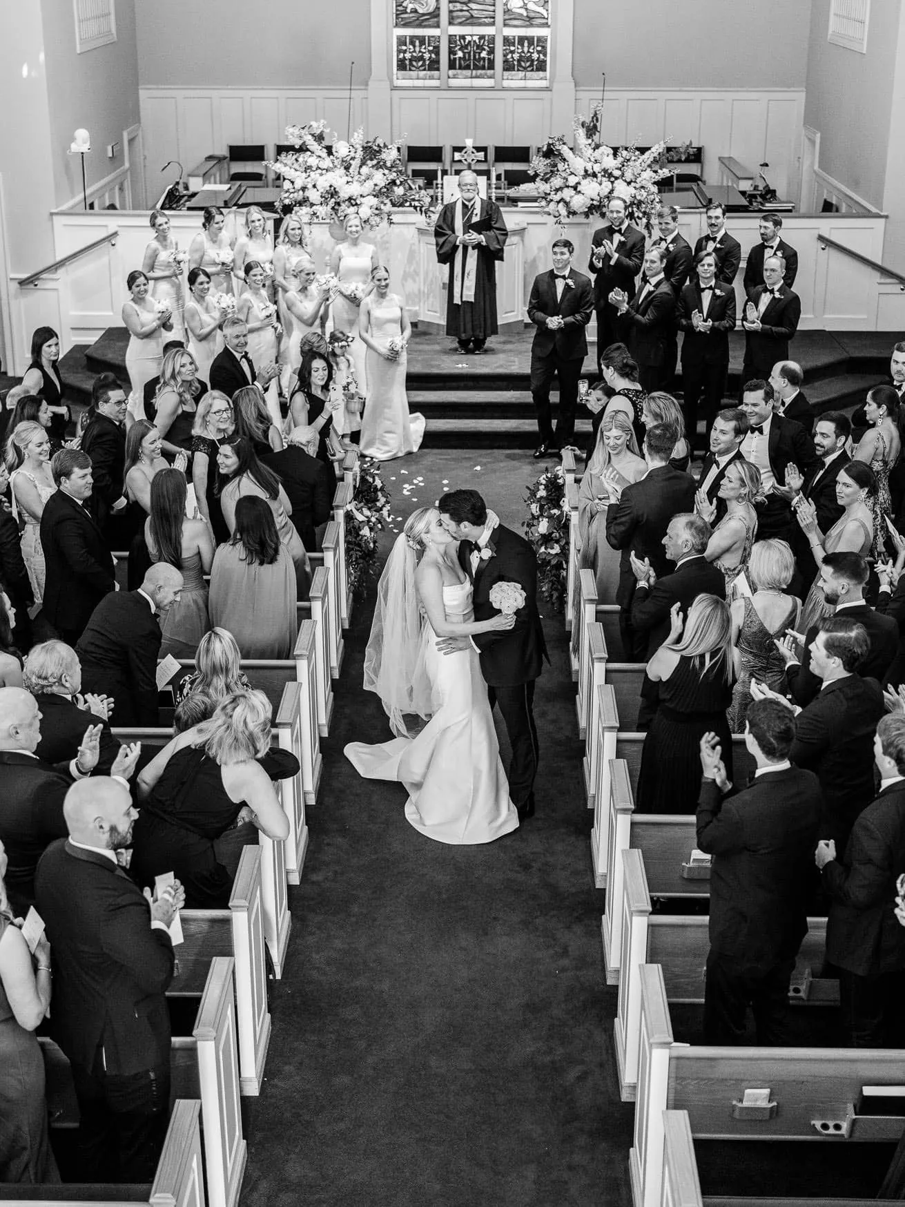 Elegant wedding ceremony captured in a church with a bride and groom sharing a kiss, surrounded by family and friends, with floral arrangements and historical stained glass windows.