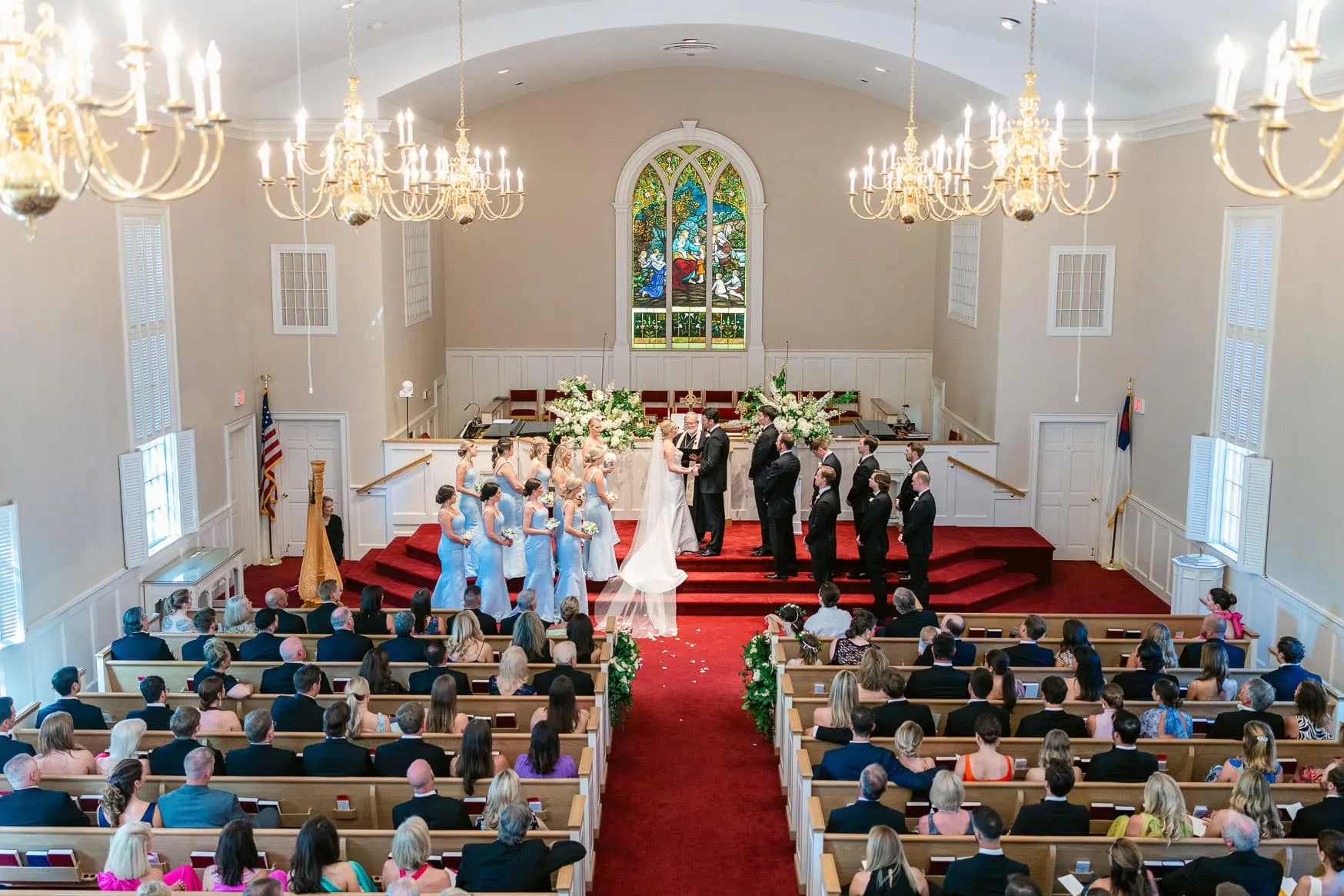 Elegant wedding ceremony inside a church with a bride and groom exchanging vows at the altar, surrounded by bridesmaids and groomsmen, stunning stained glass window in the background, and seated guests in pews.