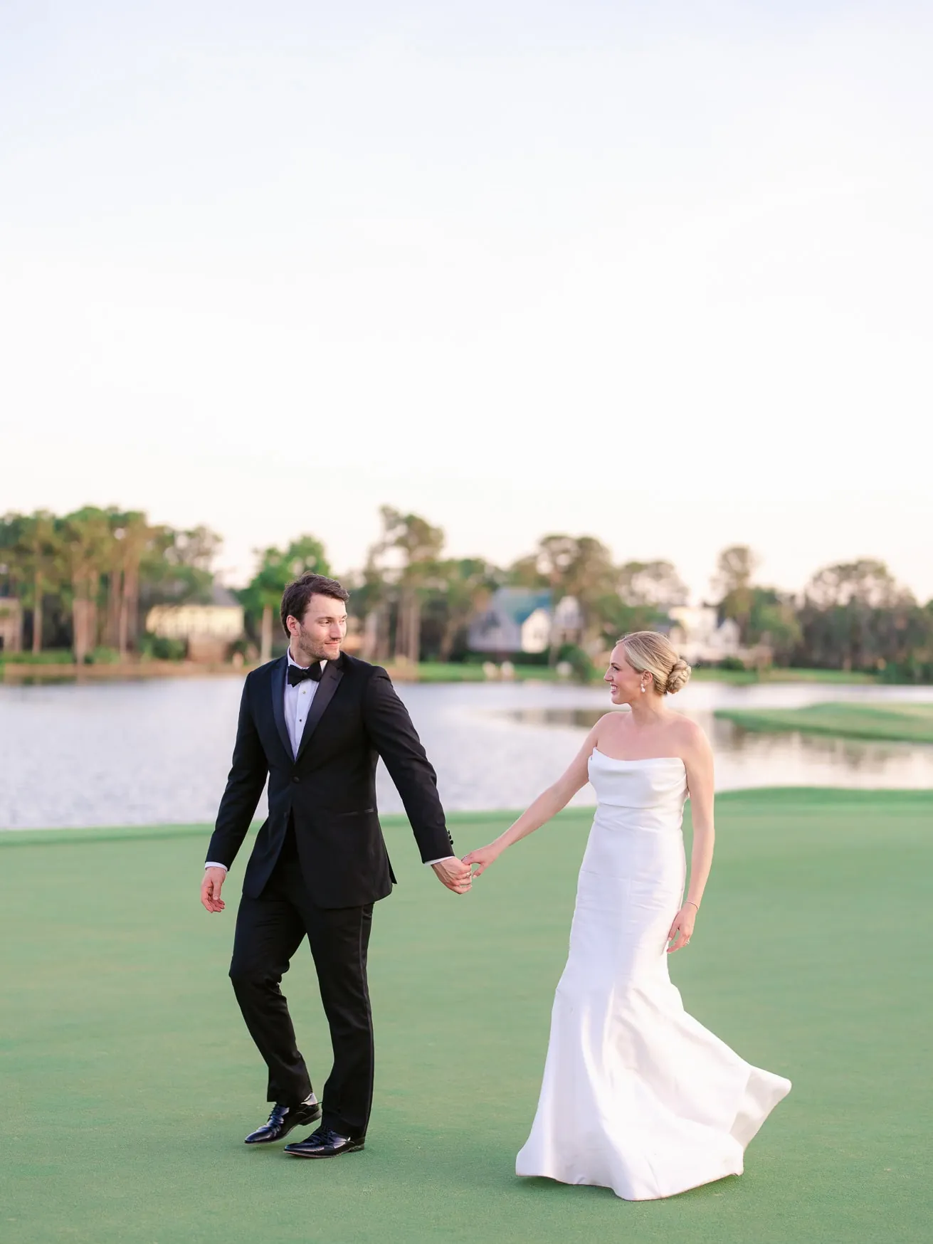 Elegant wedding couple holding hands on golf course at sunset, bridal portrait, romantic wedding photography, outdoor wedding, bride and groom, scenic lake backdrop, stylish wedding attire, joyful bride and groom, professional wedding photographer, timeless wedding moments.