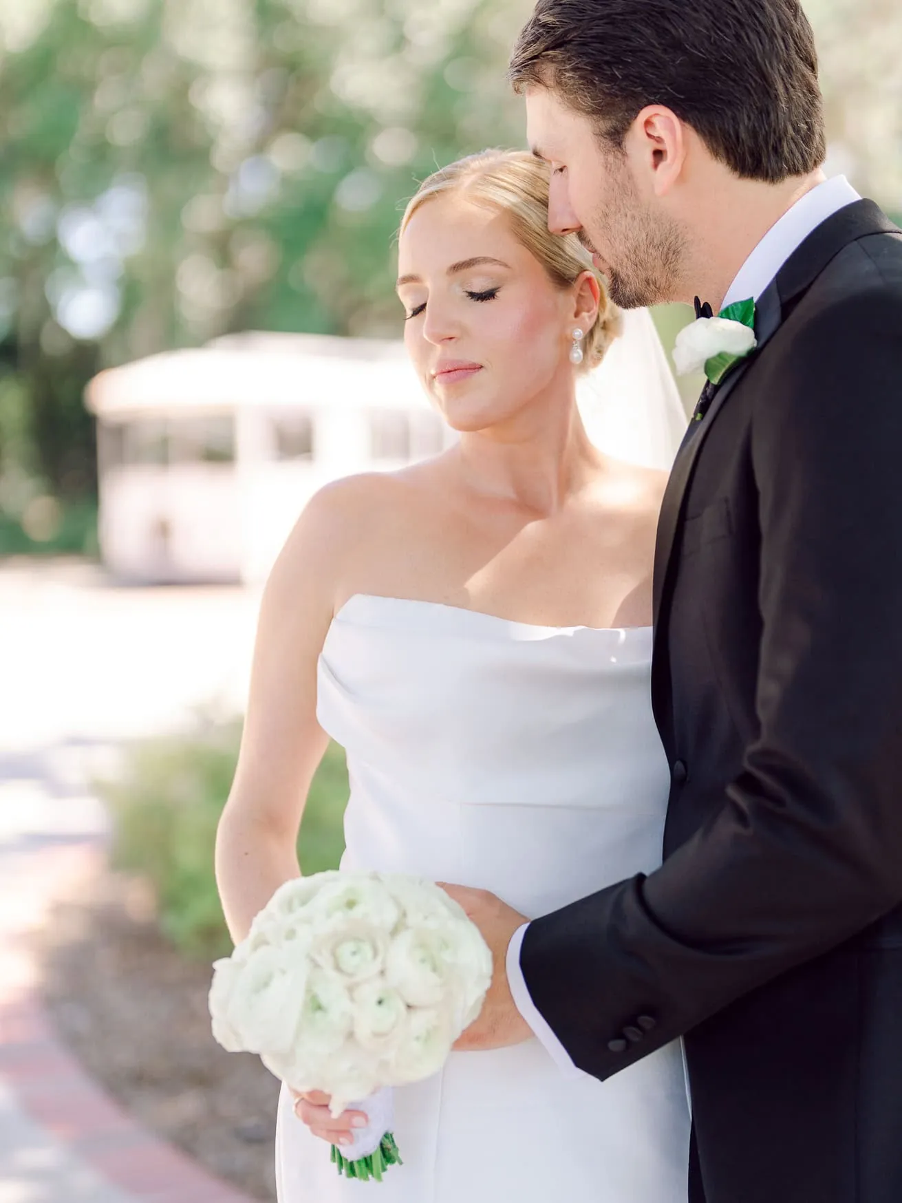 Elegant wedding couple outdoors with bride holding a white rose bouquet, groom in black tuxedo, soft natural lighting, romantic wedding portrait shot.