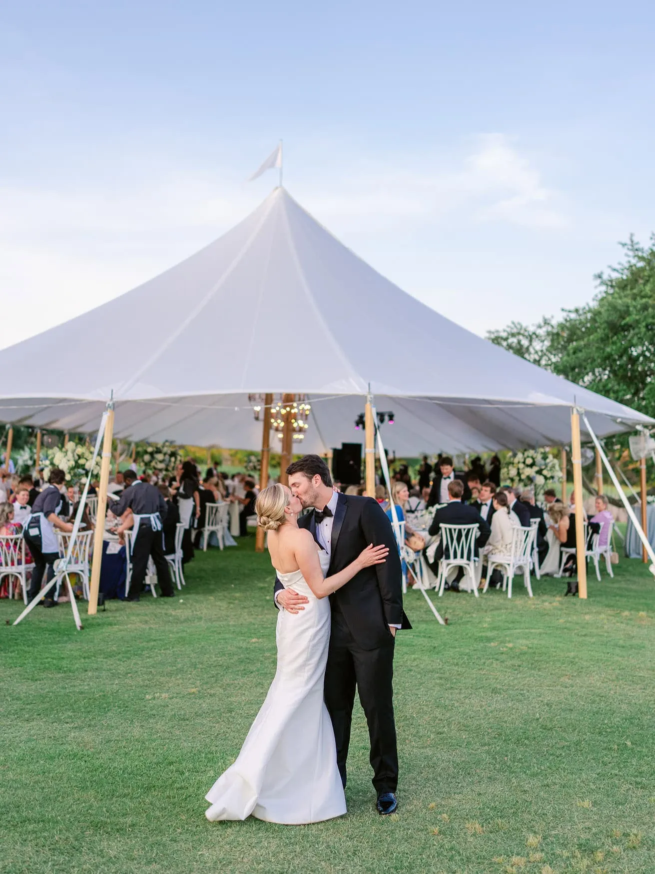 Elegant wedding couple sharing first dance under a large white wedding tent during outdoor reception, with lush greenery and softly lit party ambiance.