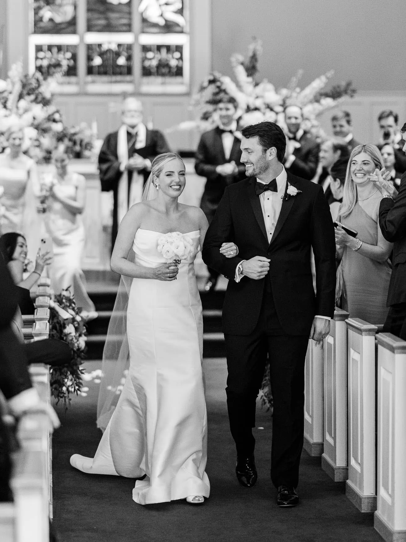 Elegant wedding couple walking down the aisle in a church, black and white photography, bride in strapless gown holding bouquet, groom in tuxedo, surrounded by joyful guests and floral arrangements.