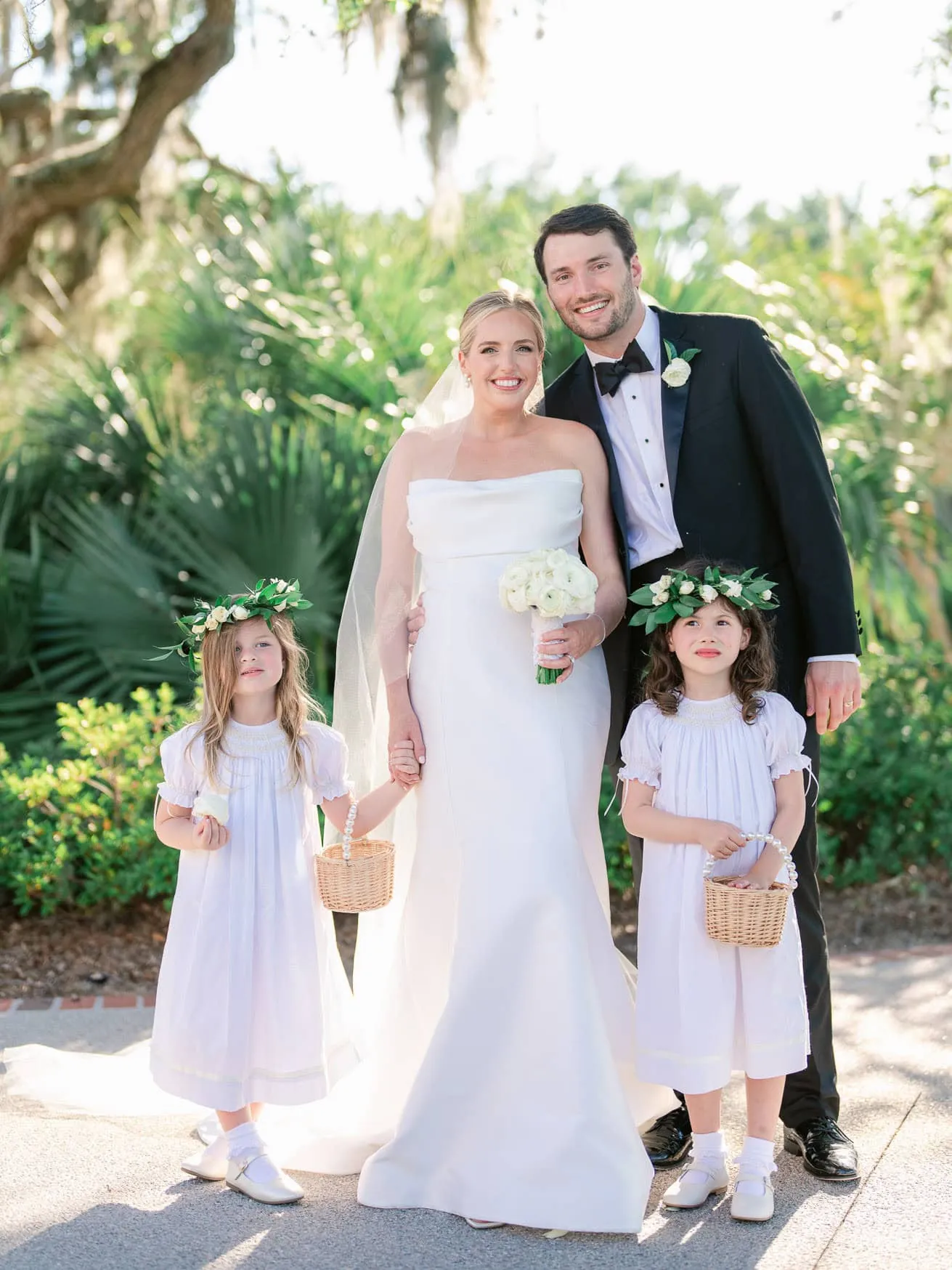 Elegant wedding portrait of a bride and groom with two young flower girls holding baskets, outdoor garden setting, natural sunlight, capturing joyful wedding moments, professional wedding photography in Florida.