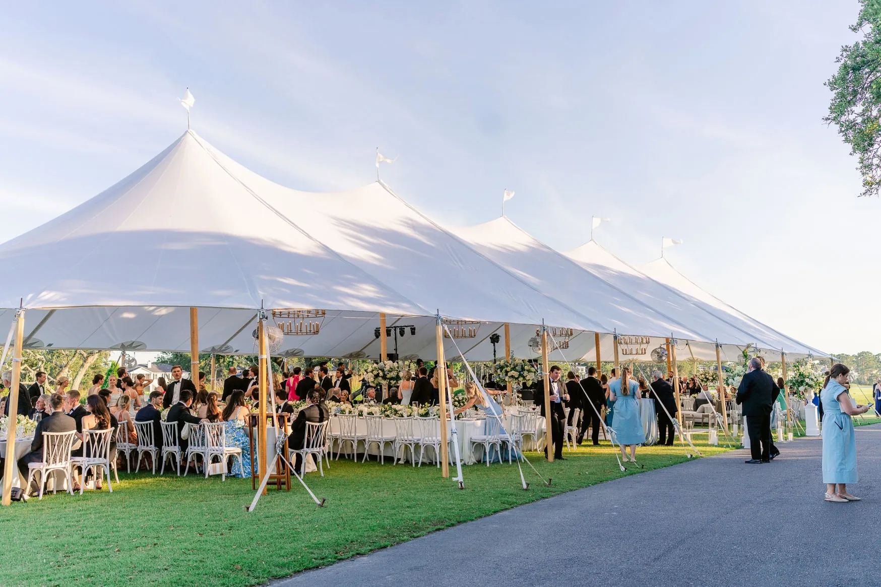 Elegant wedding reception under a large white tent with guest tables, floral arrangements, and string lights, set outdoors on a sunny day.