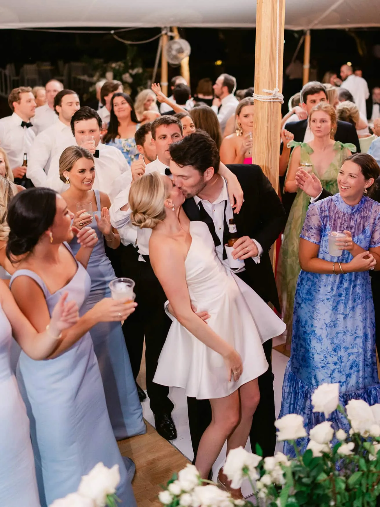 Lively wedding reception with bride and groom sharing a kiss on the dance floor, surrounded by friends and family enjoying the celebration under a tent at sunset.