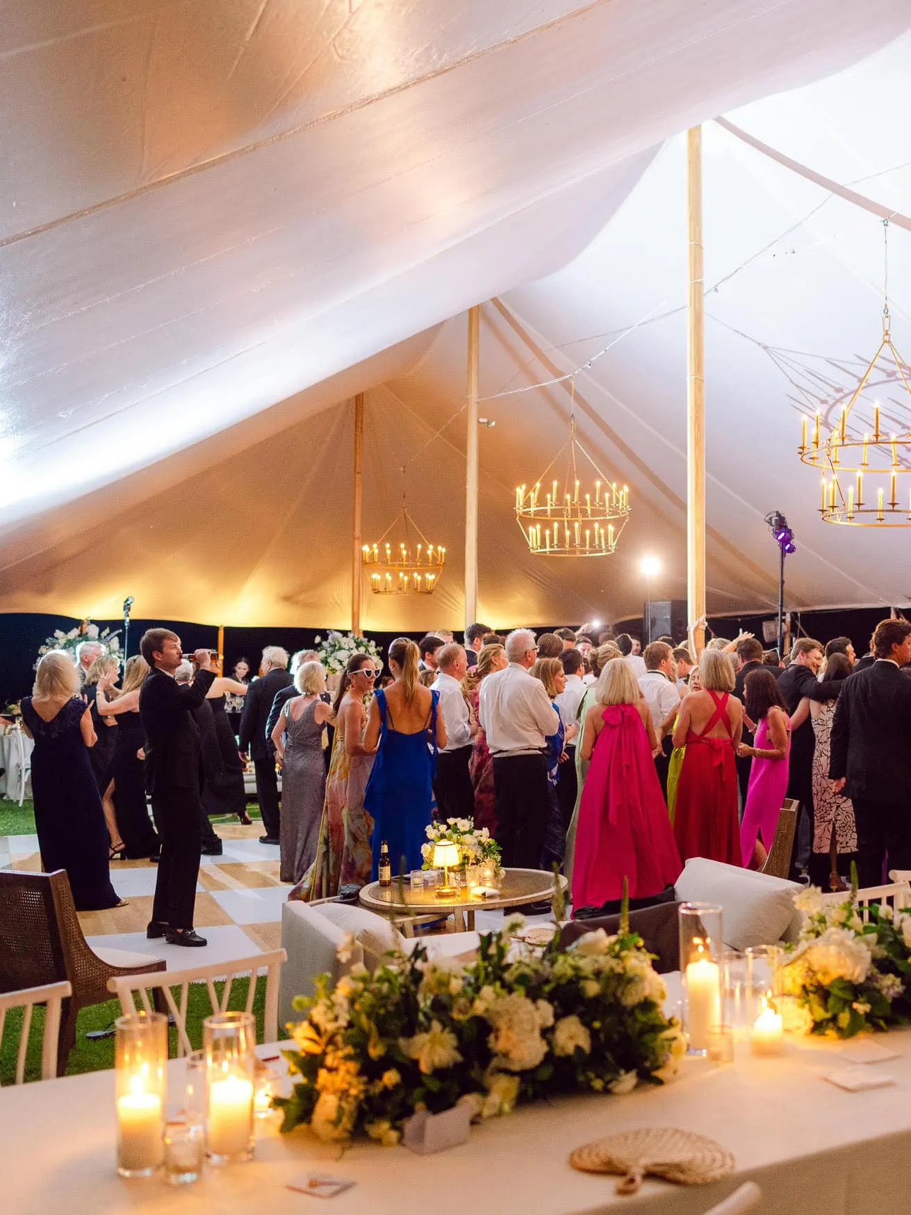 Luxurious wedding reception under a large white tent with elegant chandeliers, dim candle lighting, and guests dancing and celebrating in formal attire.