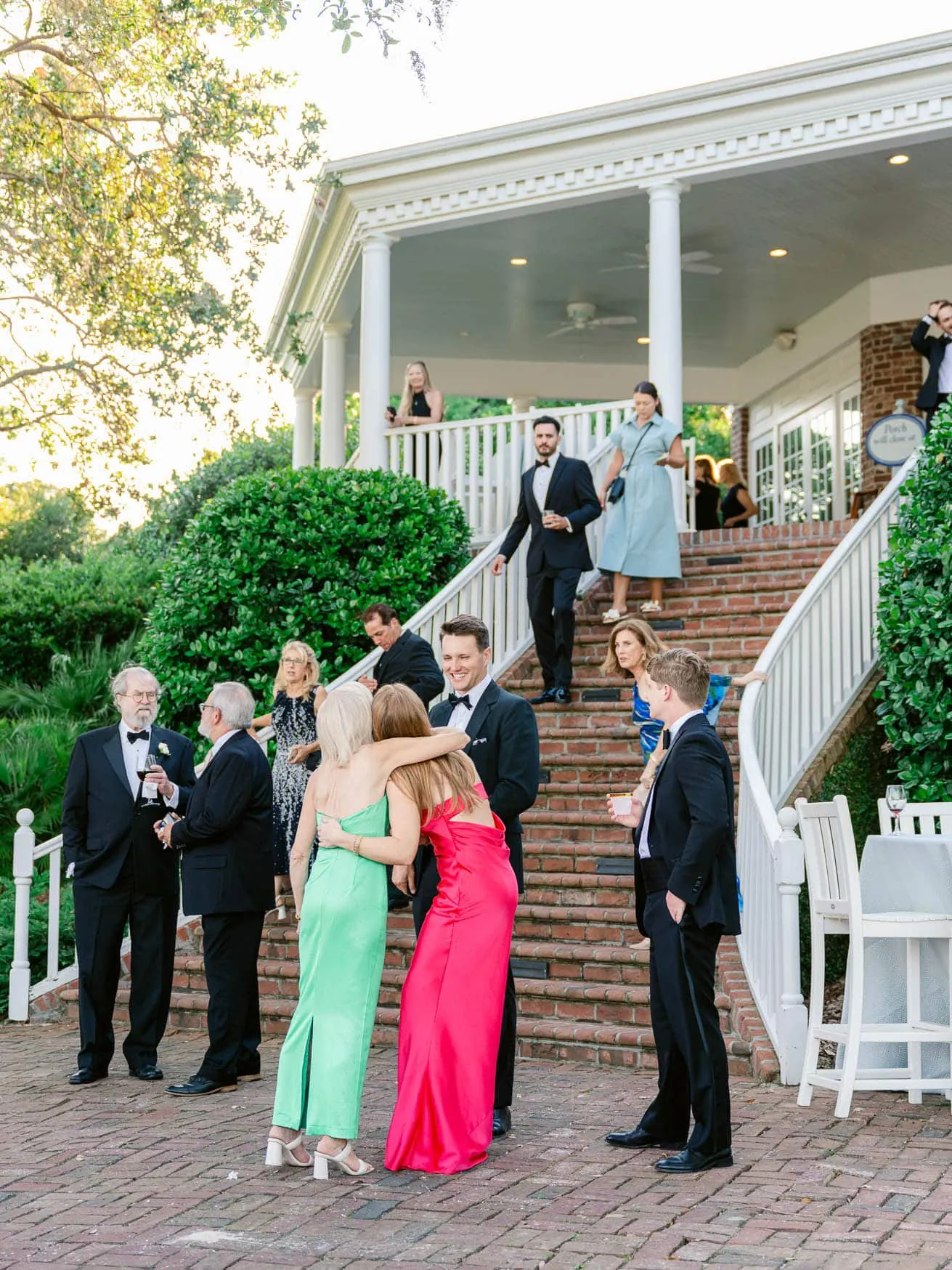 People at a formal outdoor event, socializing on a brick patio with a historic building and lush greenery in the background.