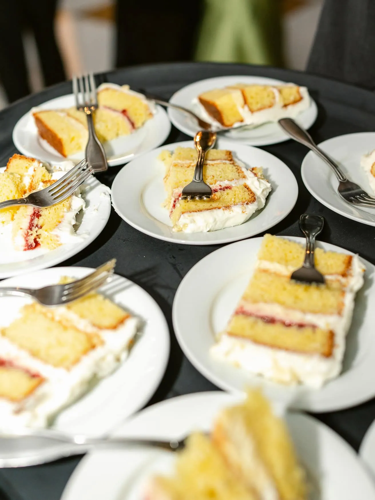 Slices of vanilla cake with cream and jam on white plates, forks resting on the desserts, served on a black tablecloth, at a celebratory event.
