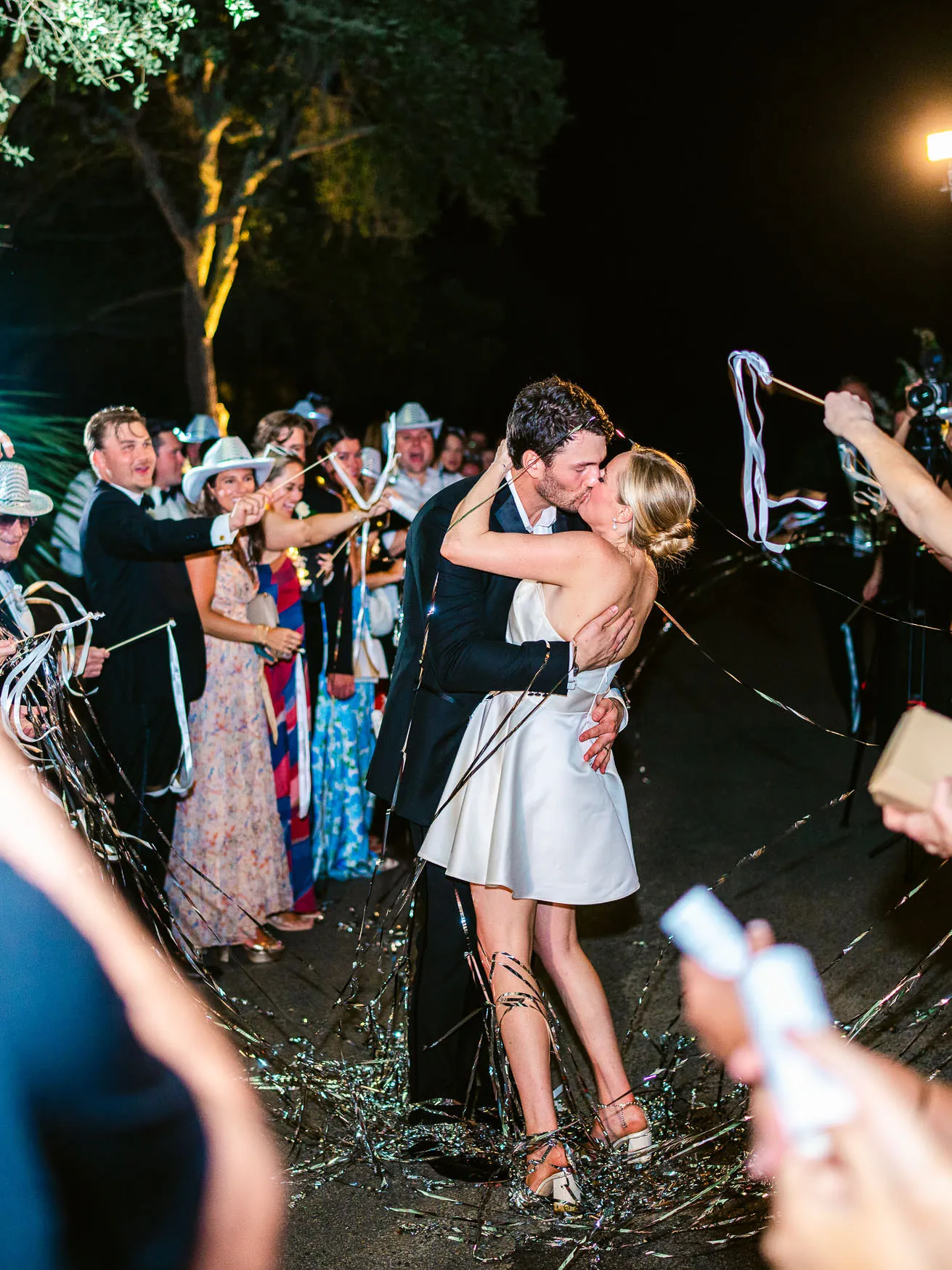 Vibrant wedding celebration scene featuring a newlywed couple sharing a kiss under nighttime lights, surrounded by friends and family in elegant attire with festive confetti decorations.