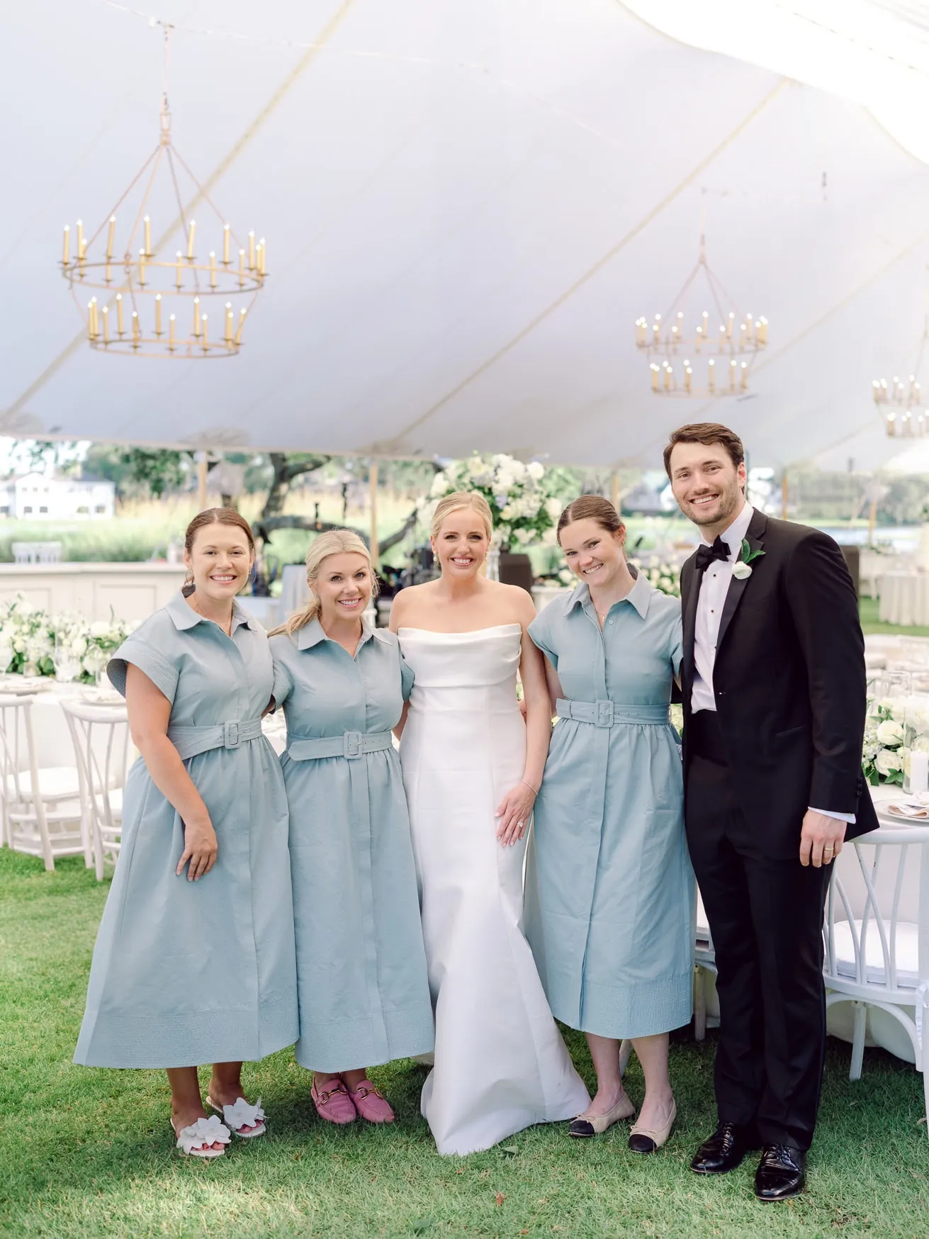 Vibrant wedding celebration with bride, groom, and bridesmaids in pastel blue dresses under a white tent with elegant chandeliers. Elegant outdoor wedding photography capturing joyful moments.