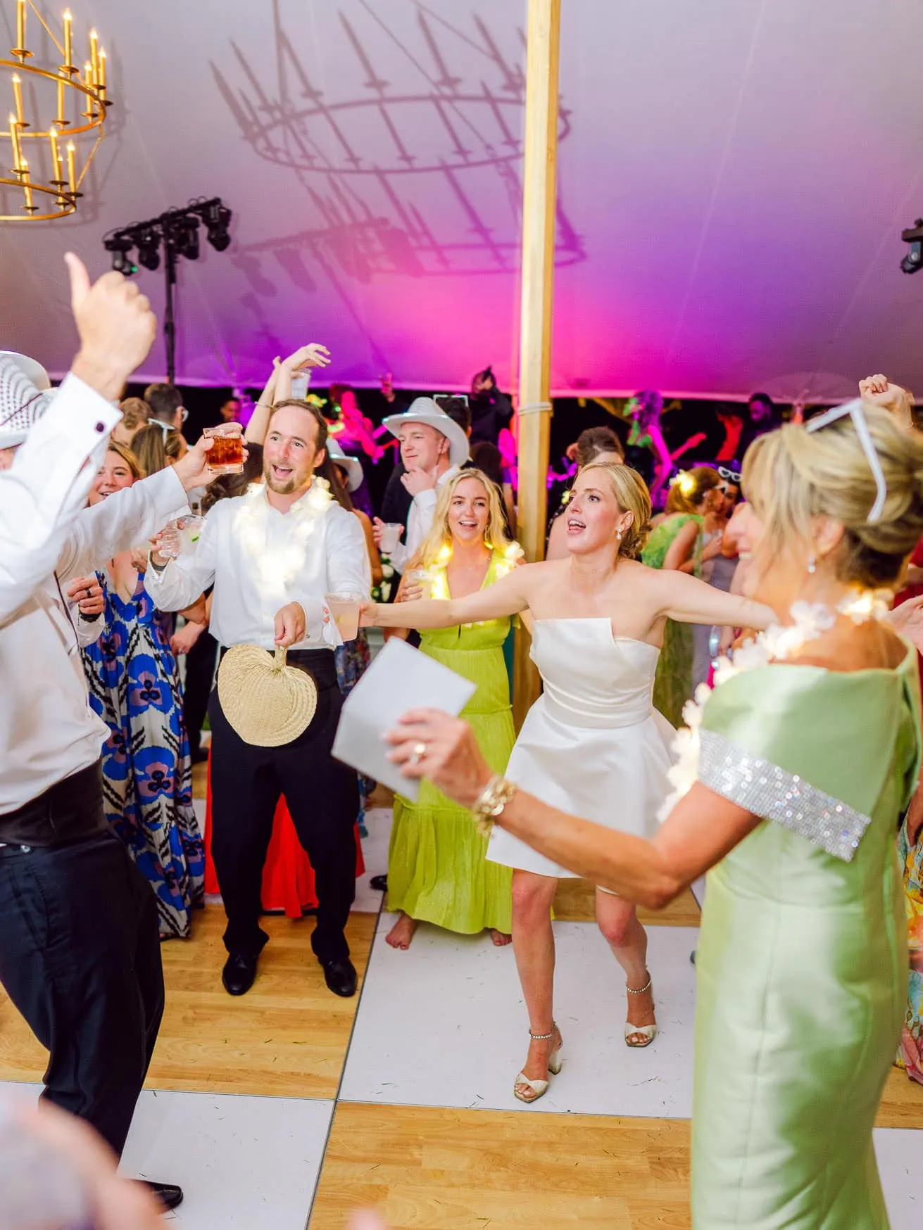 Vibrant wedding celebration with guests dancing, joyful expressions, and lively atmosphere at an indoor reception under colorful lighting.