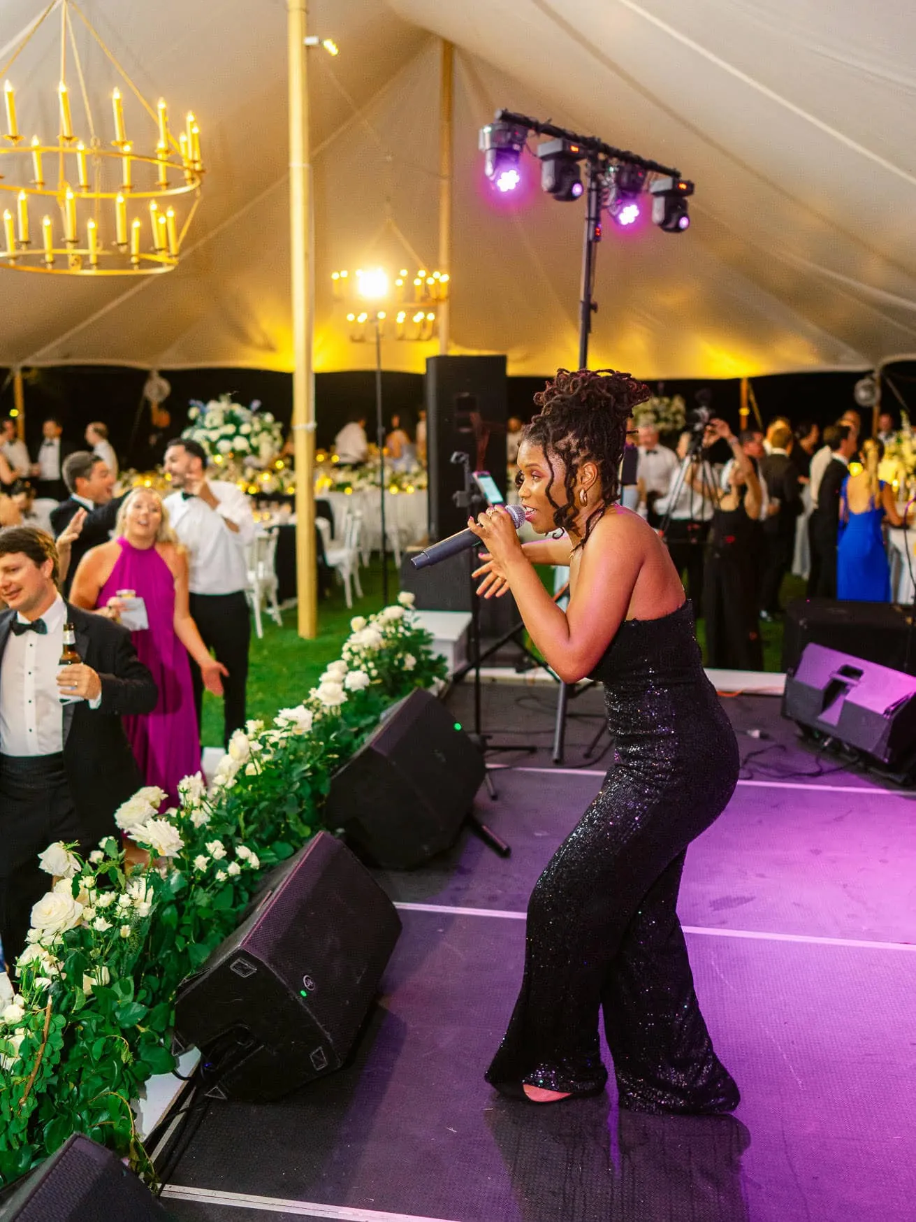 Vibrant wedding reception celebration with a female singer in a glittering black gown performing on stage under a grand chandelier in a beautifully decorated tent.