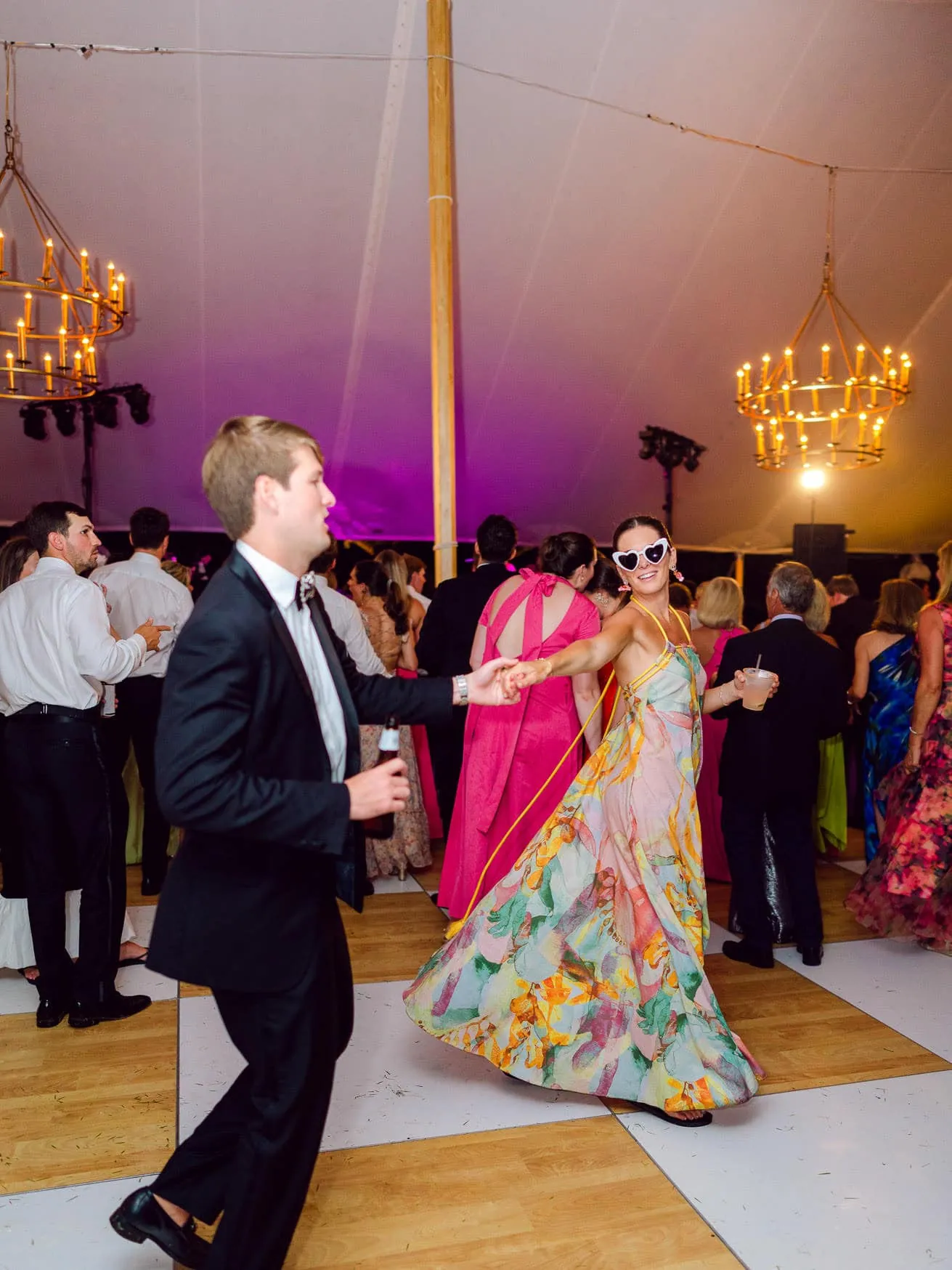 Vibrant wedding reception dance floor with joyful guests in elegant attire, colorful dress, and fun accessories, illuminated by chandelier lighting, capturing celebration and festive atmosphere.