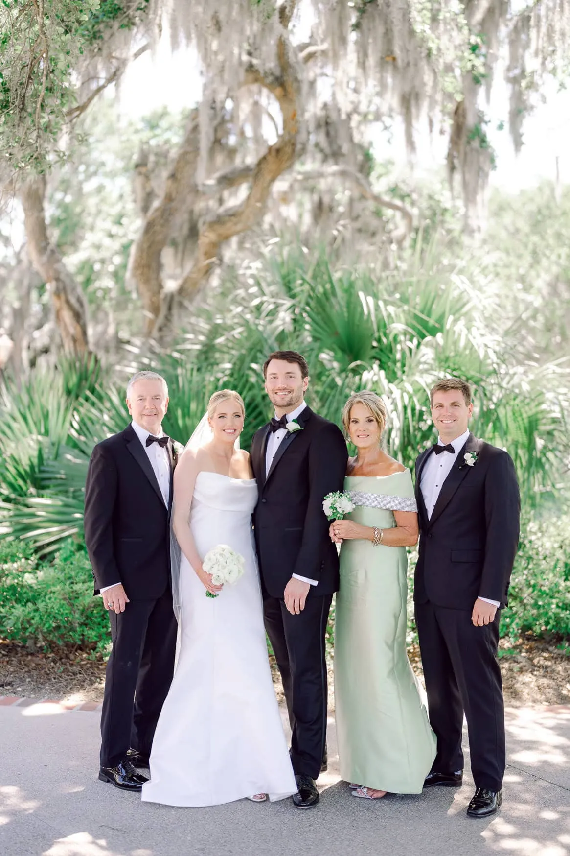 Vivid wedding portrait of a bride, groom, and family members in formal attire with lush green tropical background, capturing joyful moments of love and celebration.