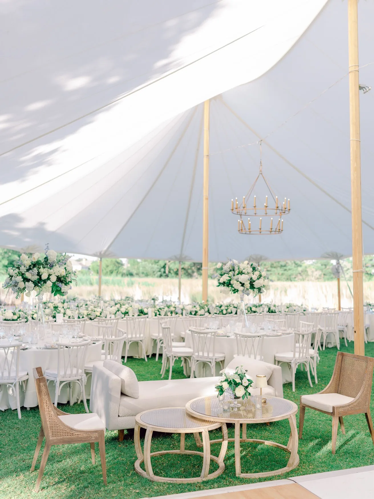 Wedding reception setup under a white tent with elegant floral centerpieces, round tables, and comfortable lounge seating on a lush green lawn.