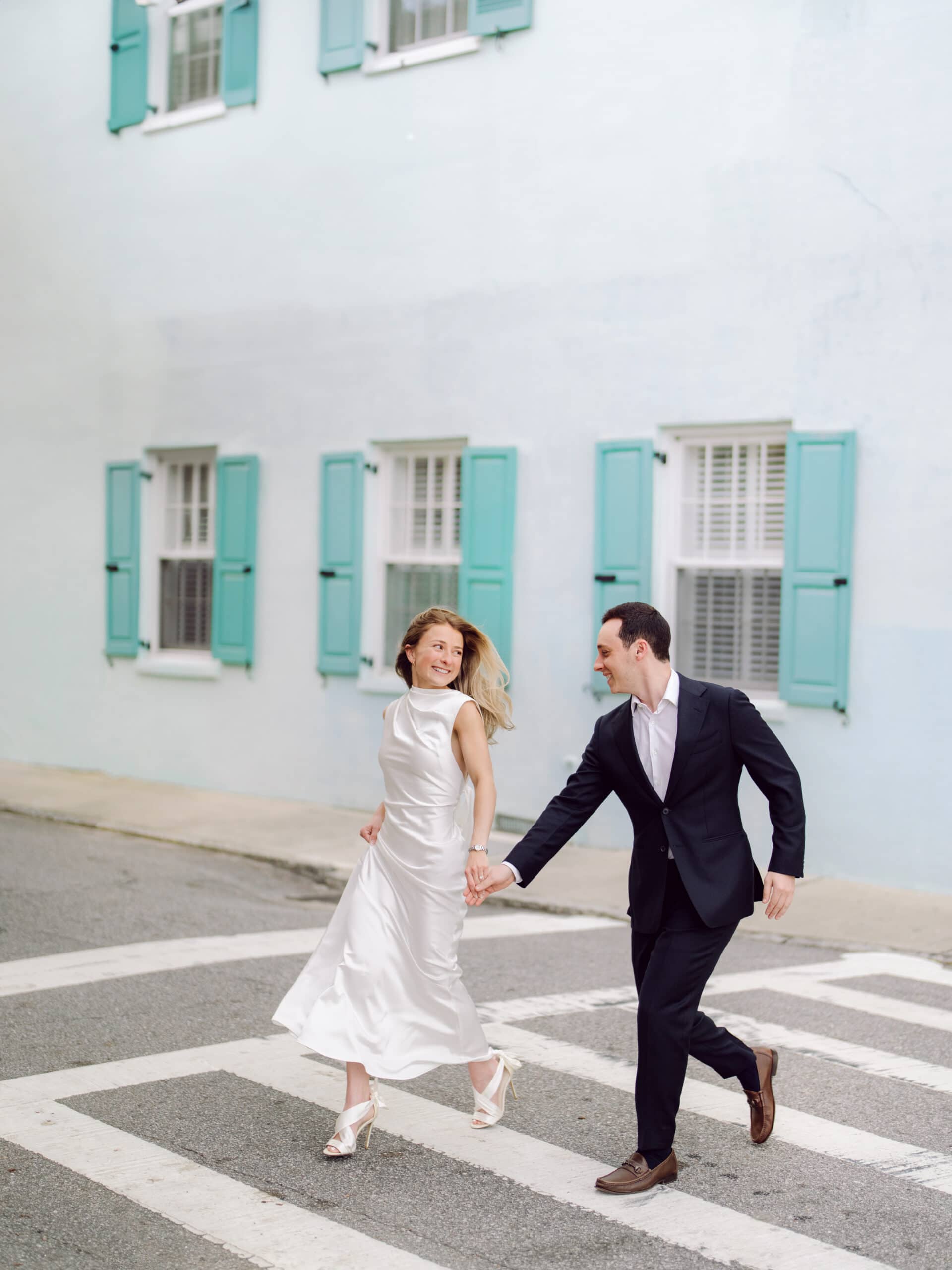 Elegant engagement photo in downtown Charleston SC with couple walking across crosswalk near pastel buildings