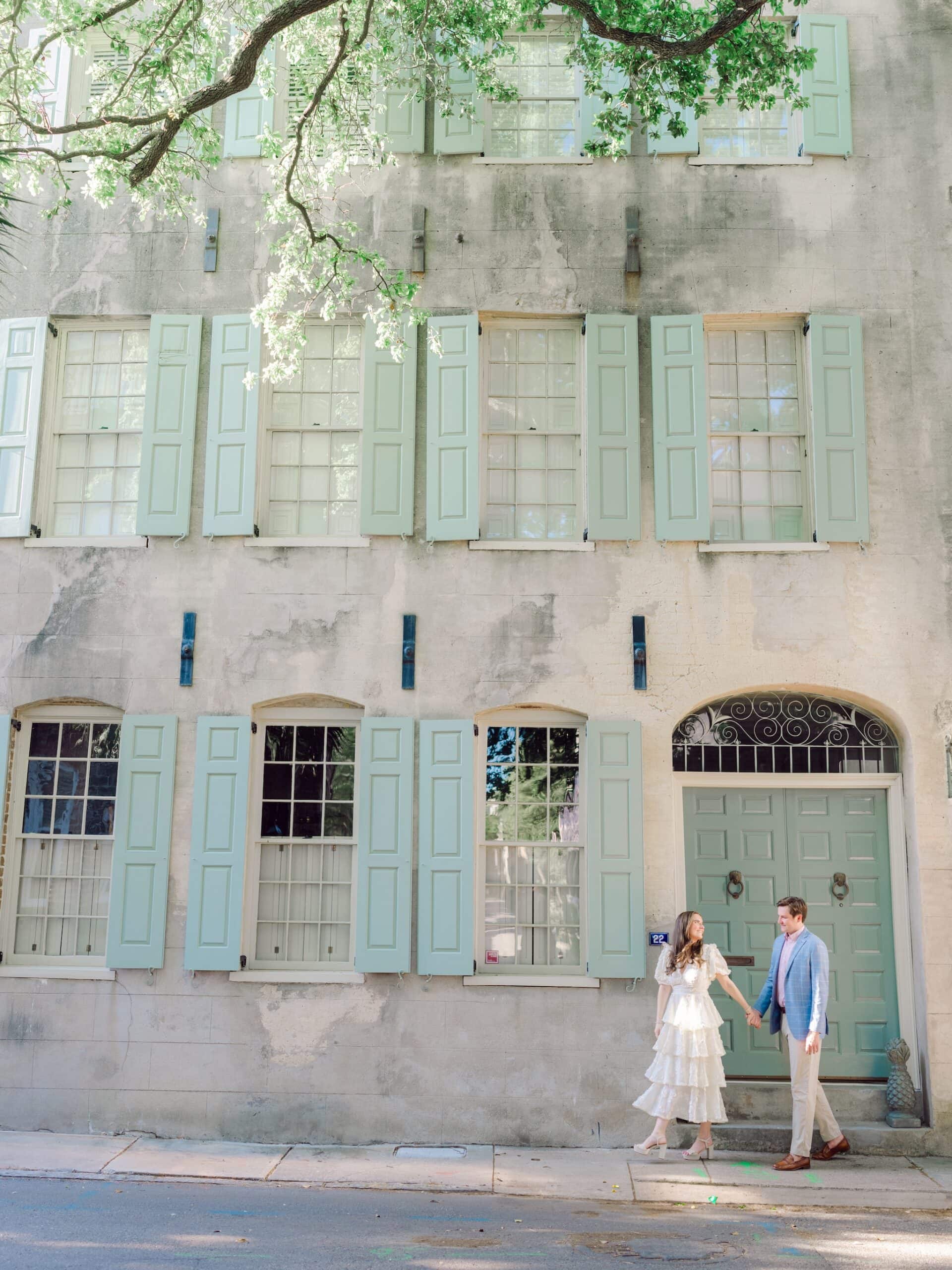 Fine art Charleston engagement portrait with couple dressed in soft tones in front of historic building with greenery