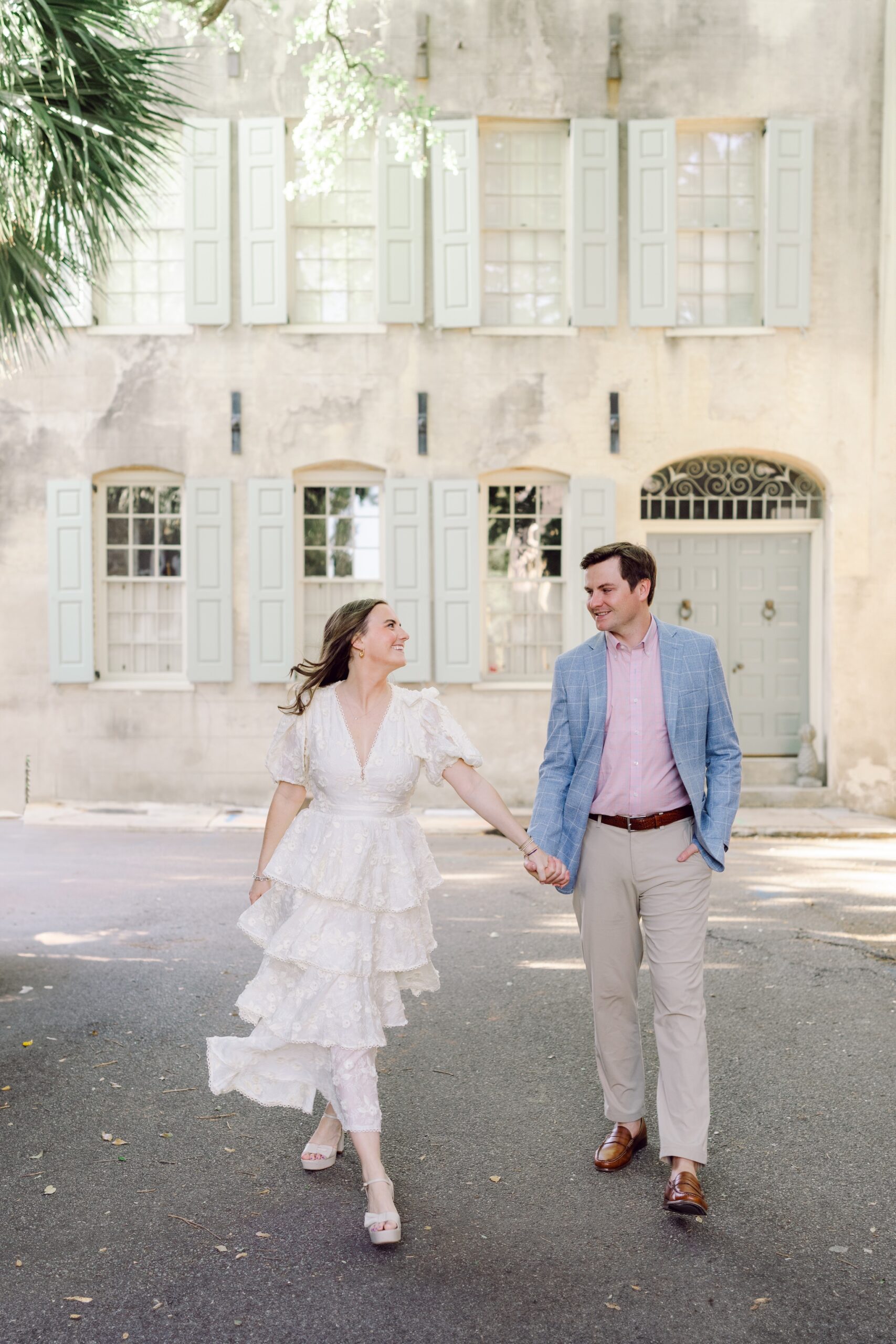 Couple holding hands during Charleston engagement shoot with charming pastel-colored historic home as backdrop