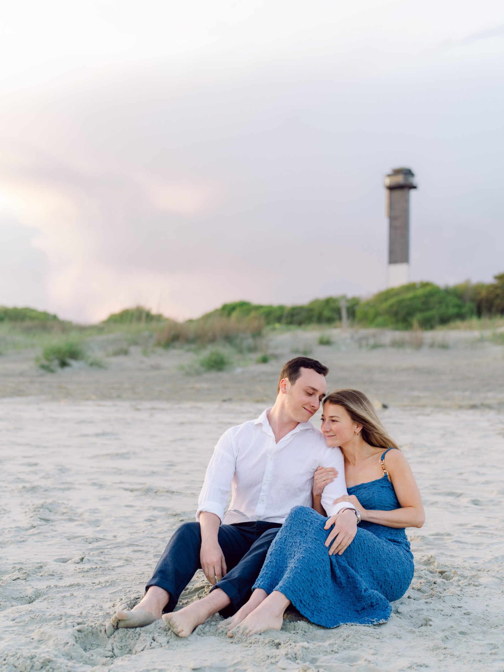 Romantic Sullivan’s Island engagement photo of couple sitting barefoot on the beach at sunset with Charleston lighthouse in background