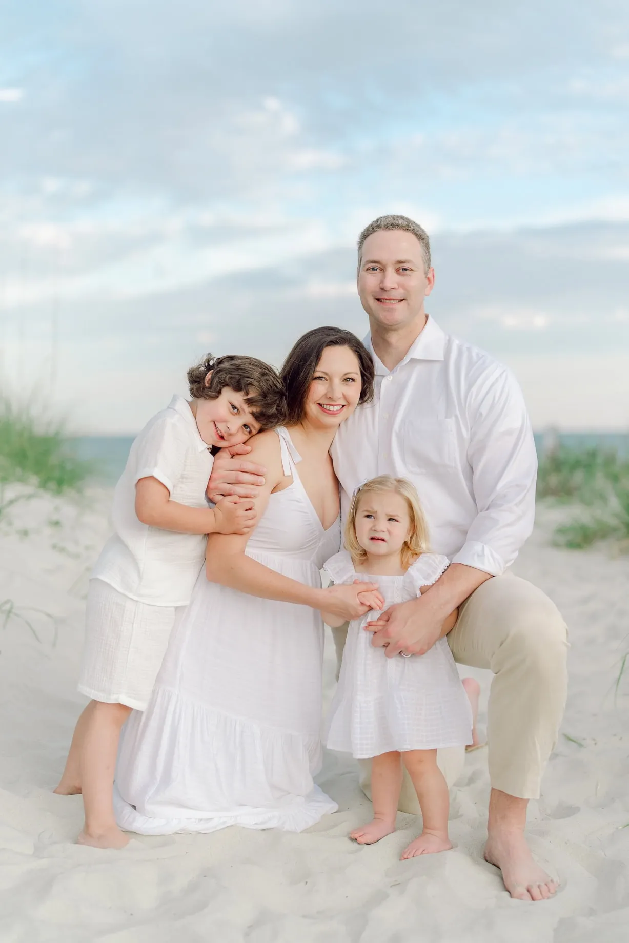 Family of four smiling and embracing during a beach photo session at Pawleys Island, with parents and two young children dressed in white, standing in soft dune grass at sunset.