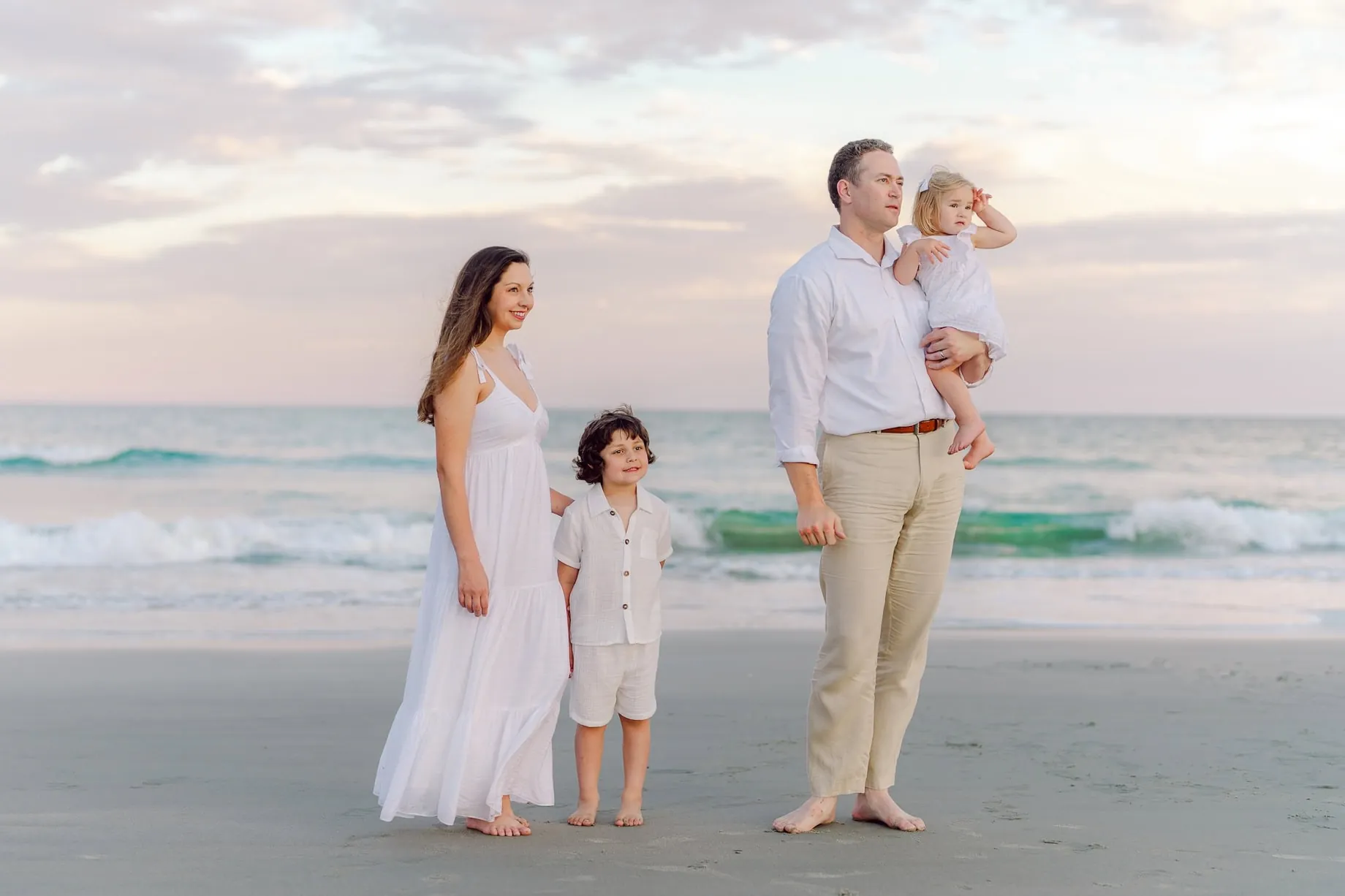 Family of four standing barefoot along the shoreline during a sunset photo session at Pawleys Island, with soft waves and pastel skies in the background.