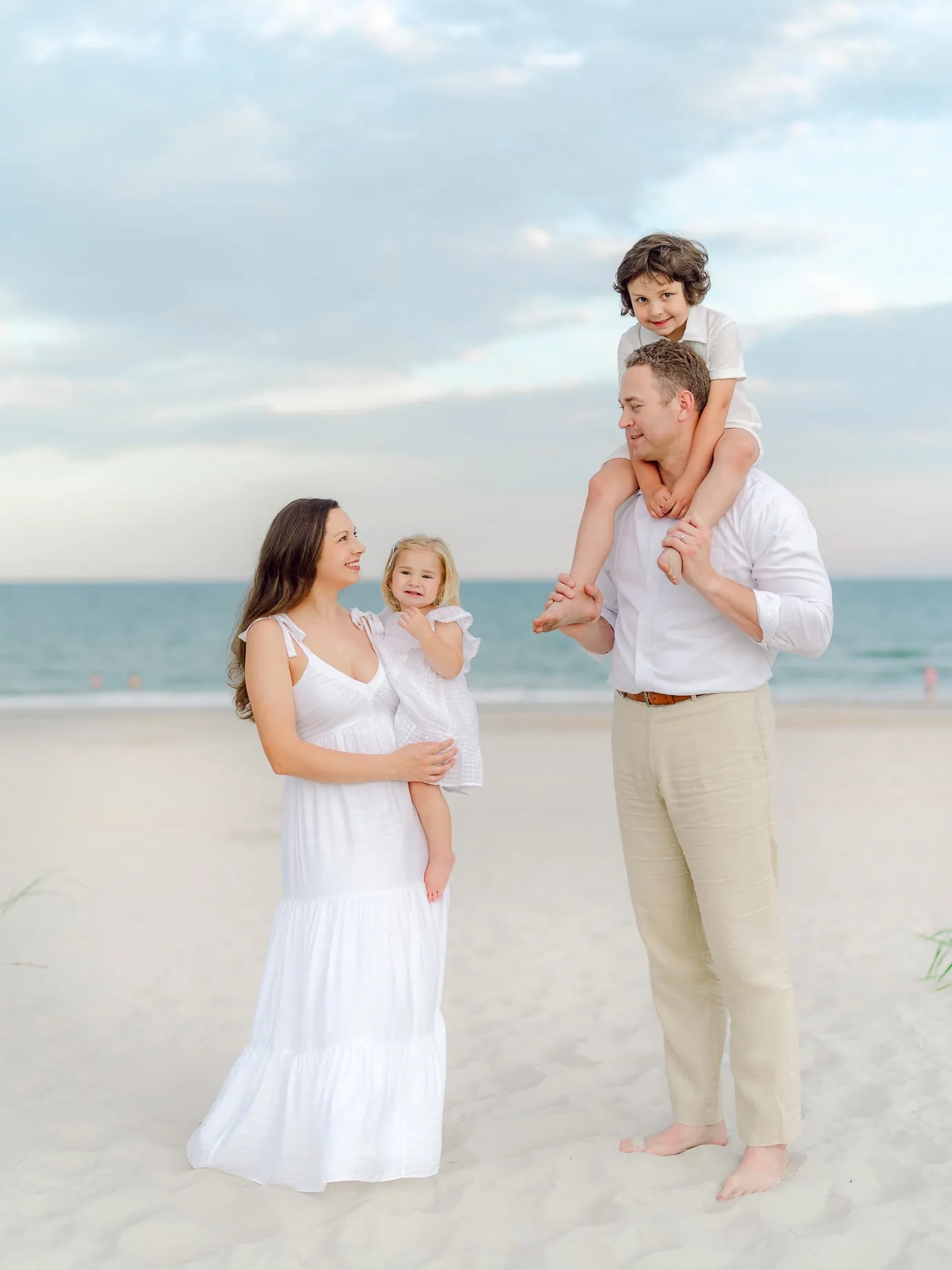 Smiling family of four during a Pawleys Island beach photo session, with the father holding a young boy on his shoulders and the mother holding a toddler daughter, all dressed in white.