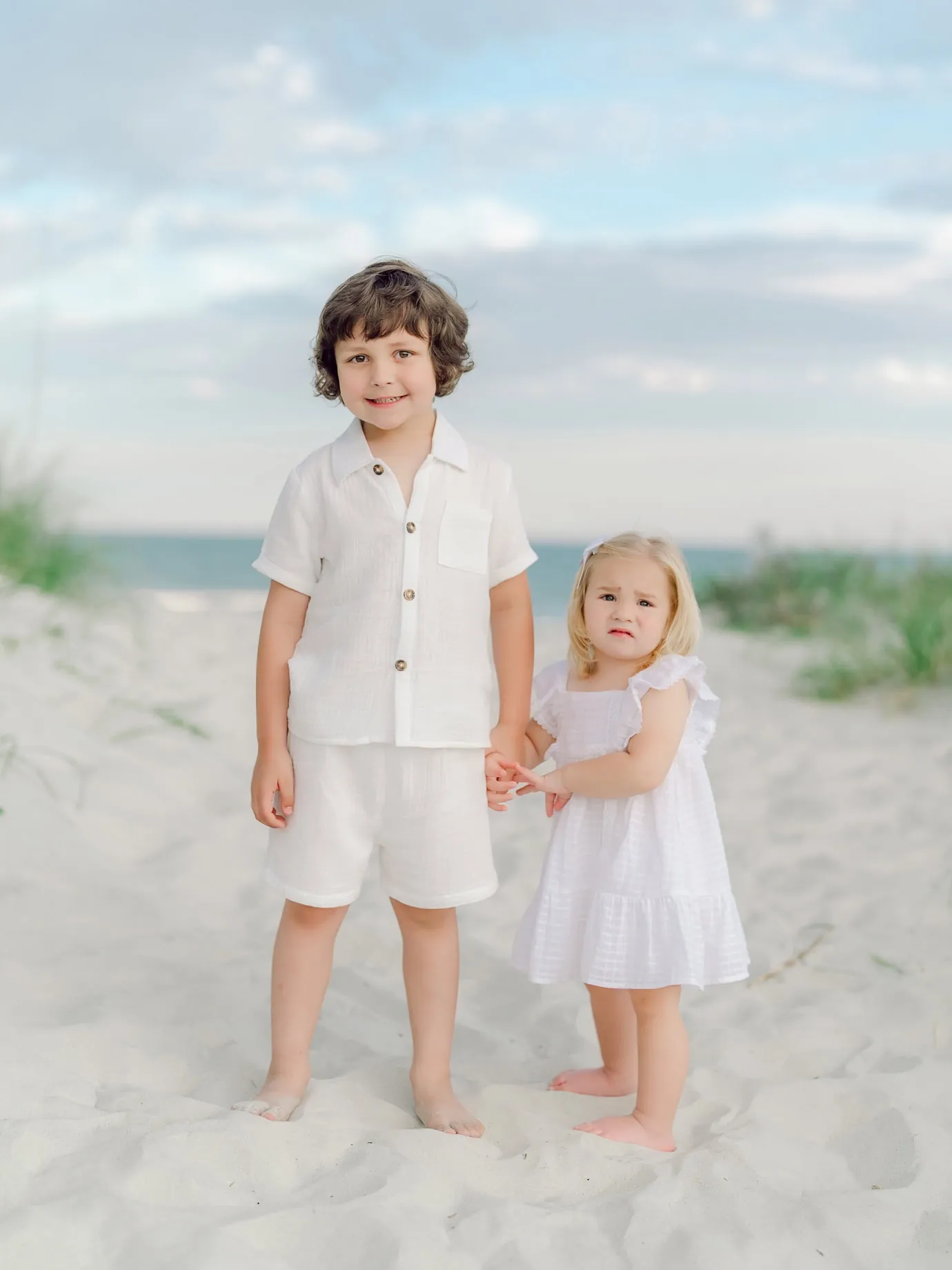 Young brother and sister holding hands during a Pawleys Island beach photo session, standing barefoot in white outfits on the sand dunes at sunset.
